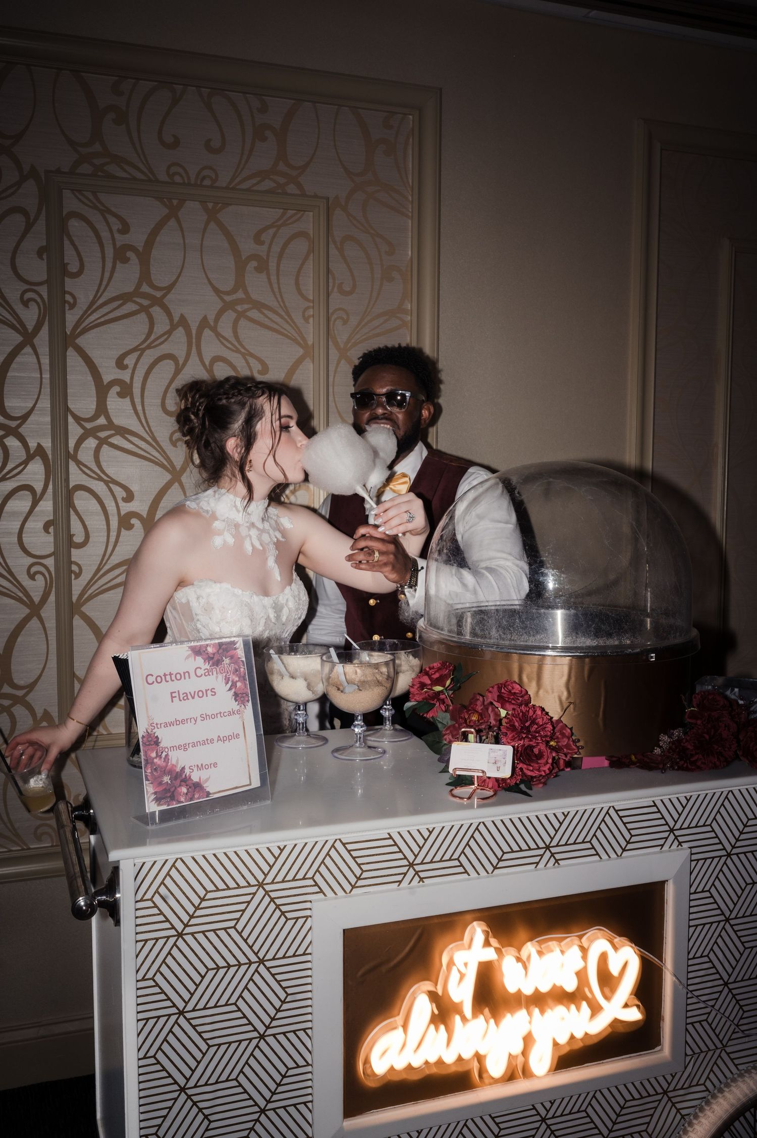 Wedding dessert display table with illuminated sign and decorative treats arranged on a patterned white surface.