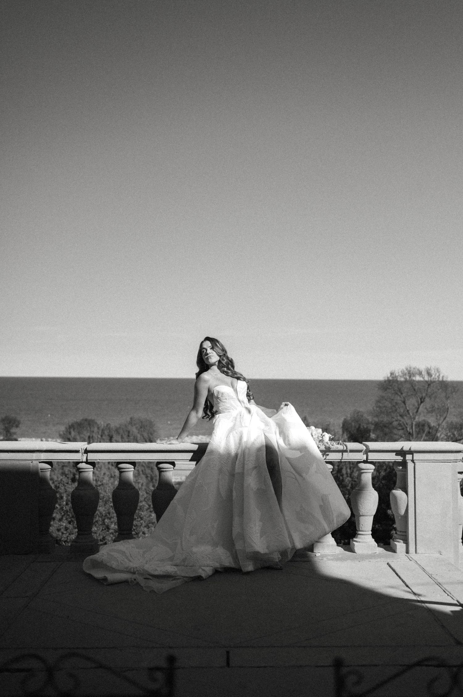 Black and white photo of bride in ballgown posing on balcony with ocean horizon in background.