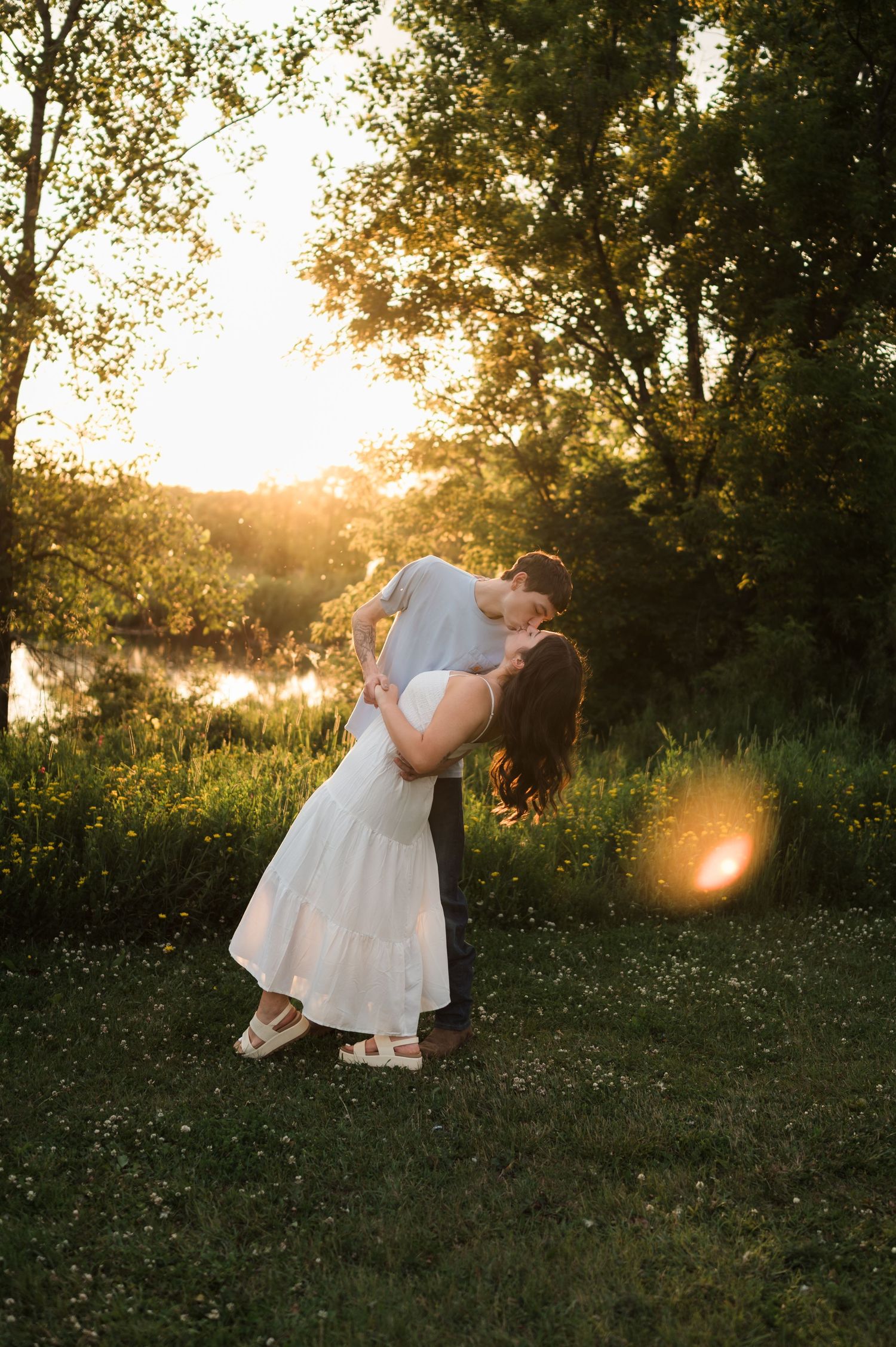 Couple sharing a romantic dip kiss in a sunlit meadow with golden hour light reflecting off a nearby pond.