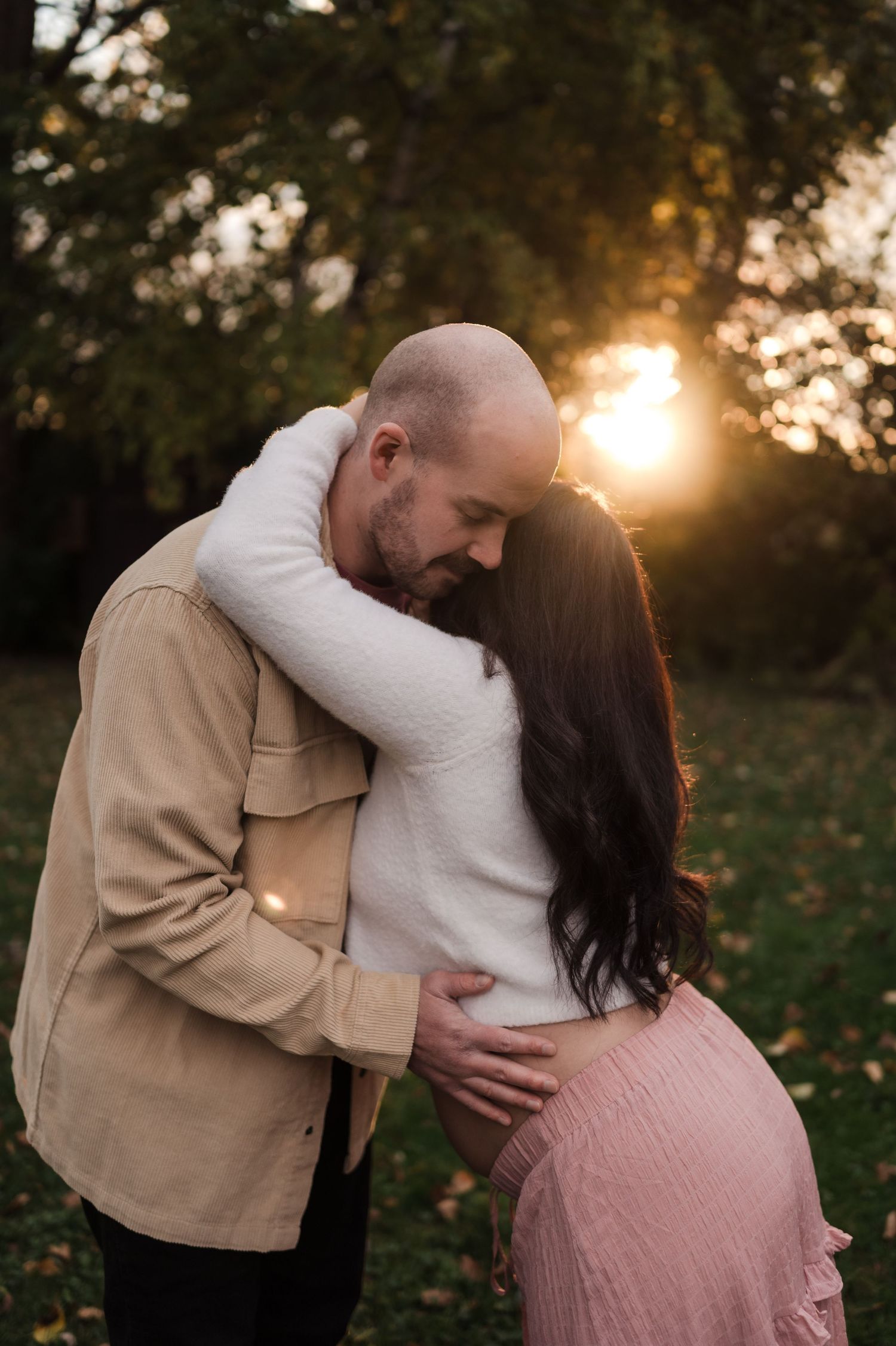 Couple sharing tender sunset embrace outdoors, man kissing woman's head while holding her baby bump.