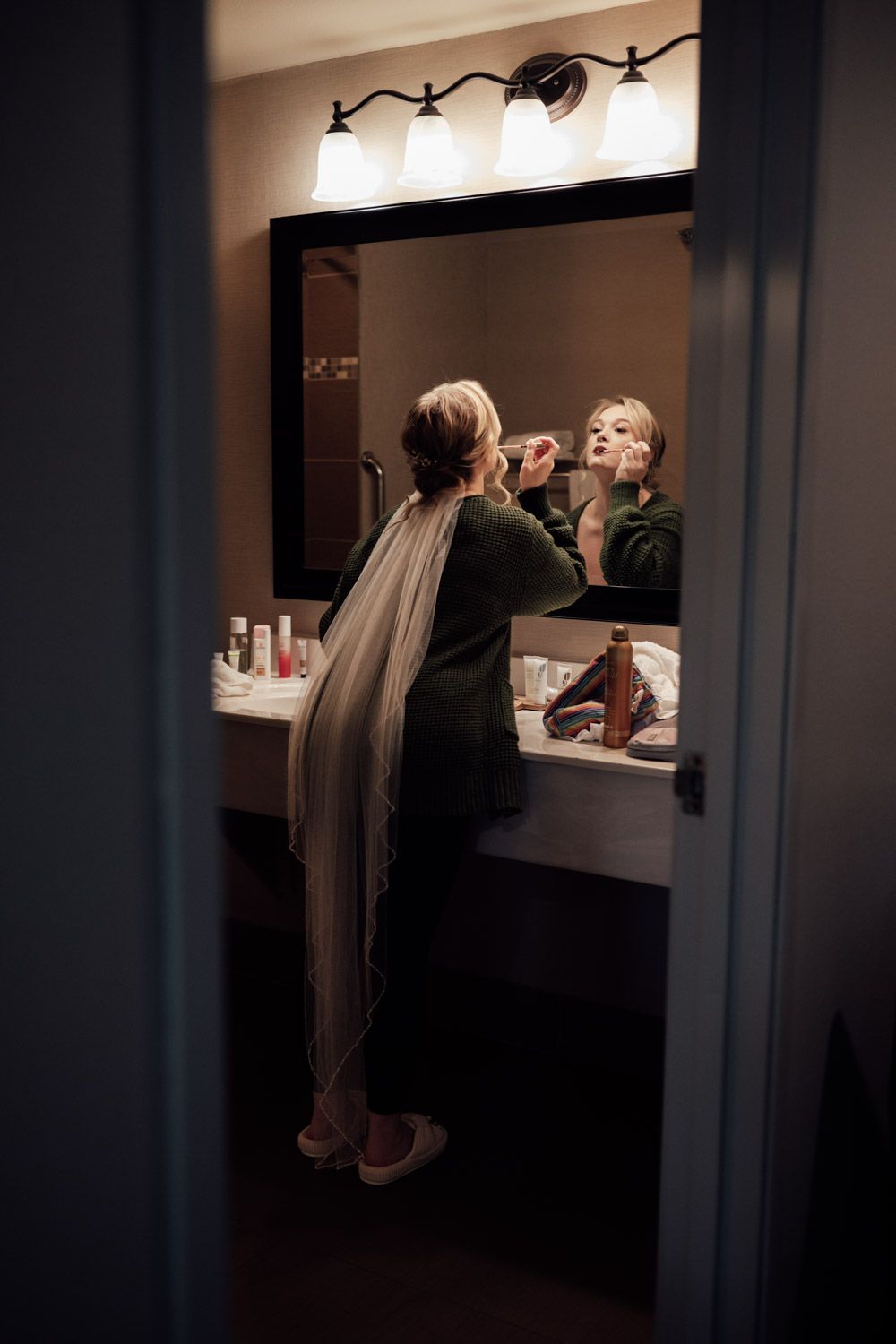 Bride in veil applying makeup at bathroom mirror while getting ready for her wedding day.