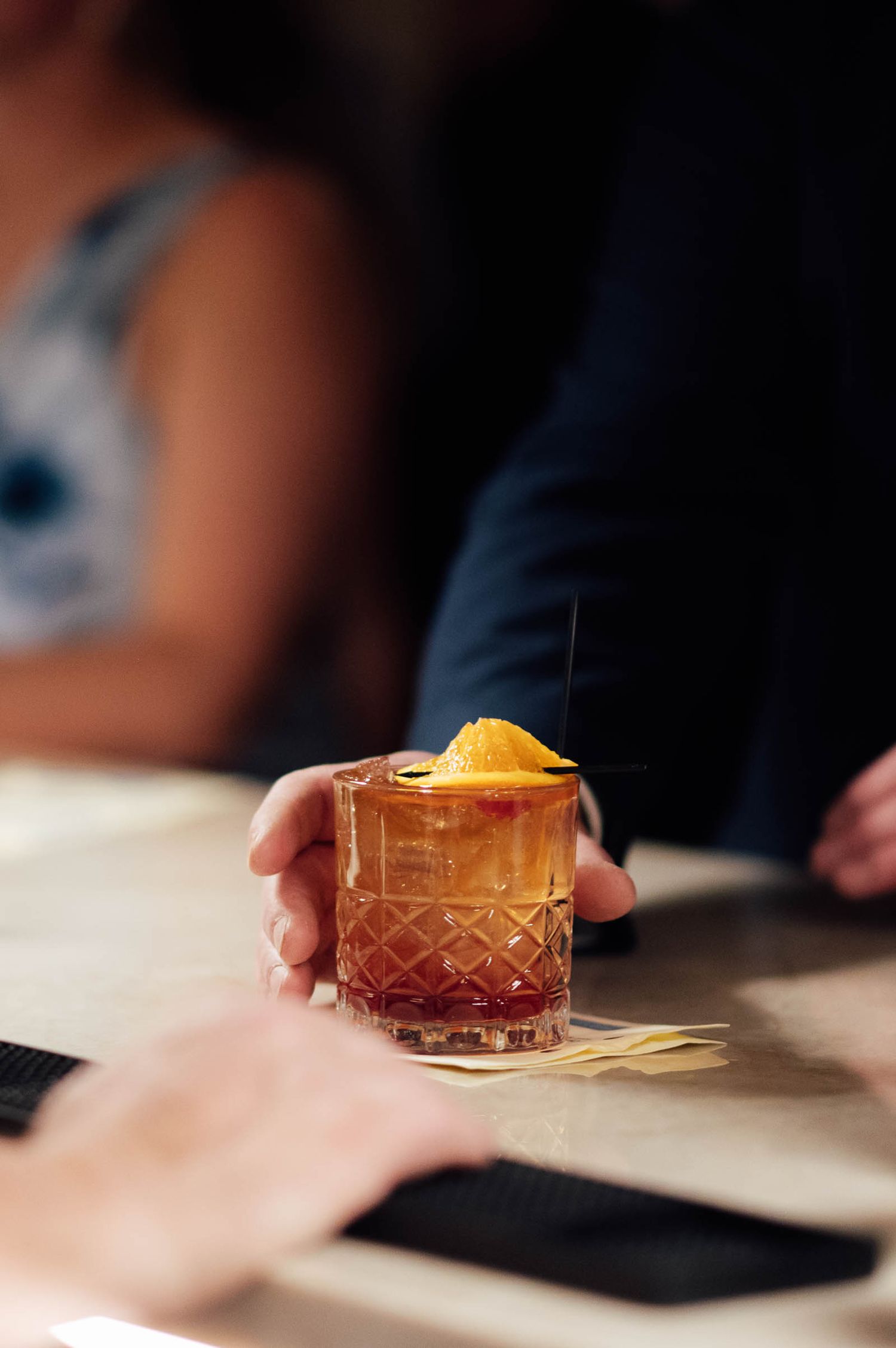 Hand holding an amber cocktail in crystal glass garnished with orange peel at wedding reception bar.