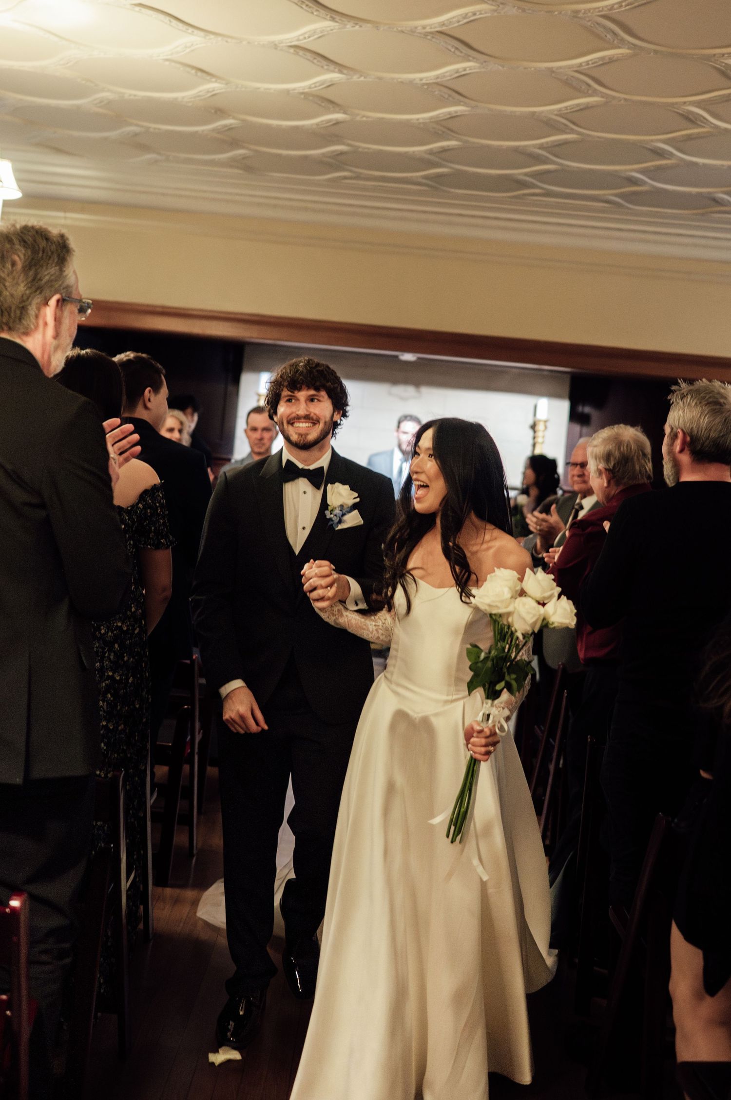 Joyful newlyweds walk down aisle hand in hand, bride holding white bouquet, guests applauding.