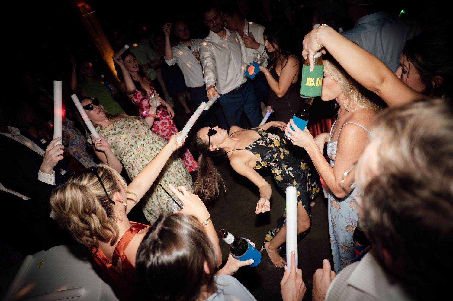 Lively wedding reception dance floor with guests holding glow sticks and drinks, celebrating at a night party.
