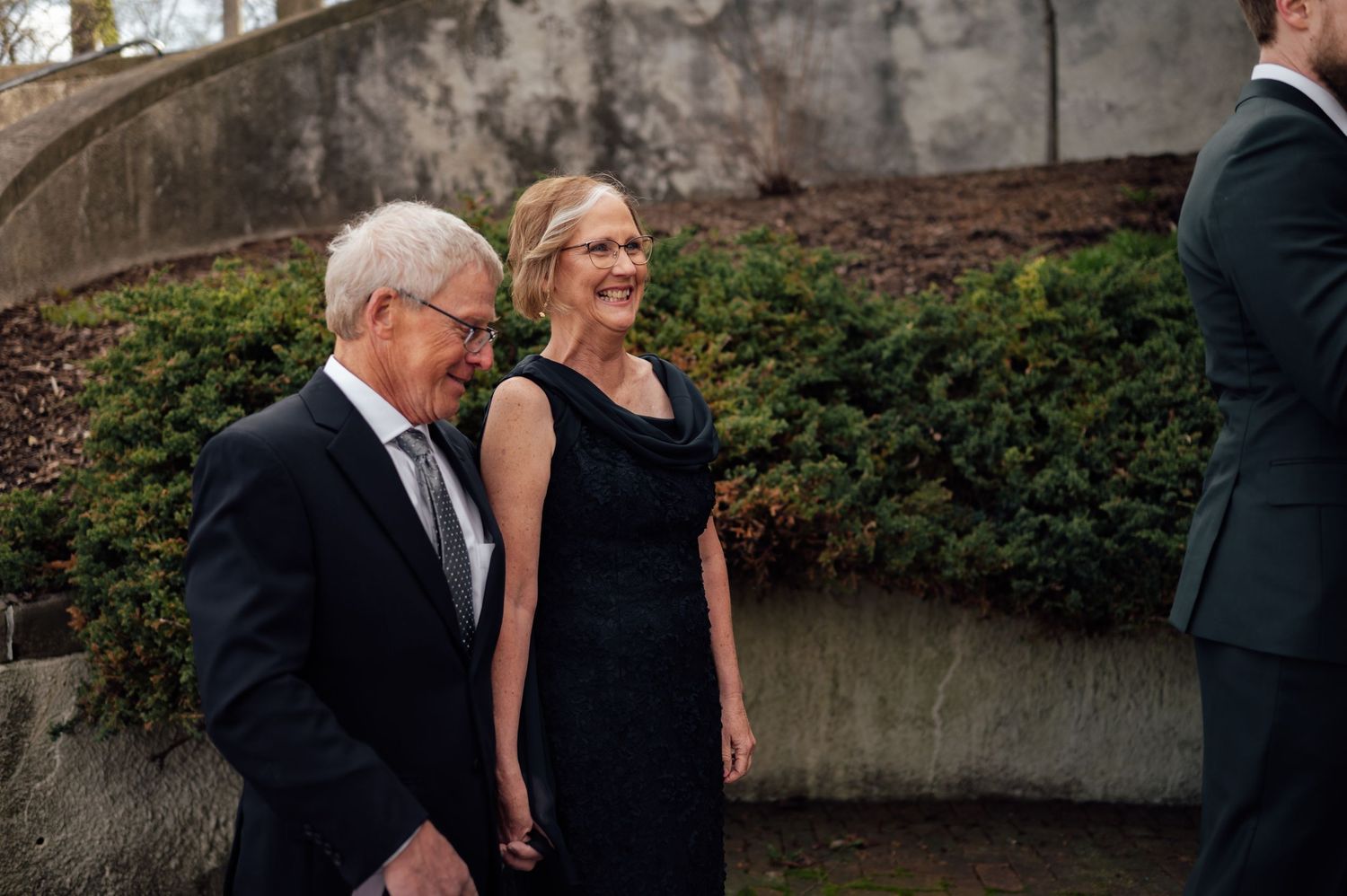 An elegant older couple in formal black attire smiling and walking together outdoors at a garden event.