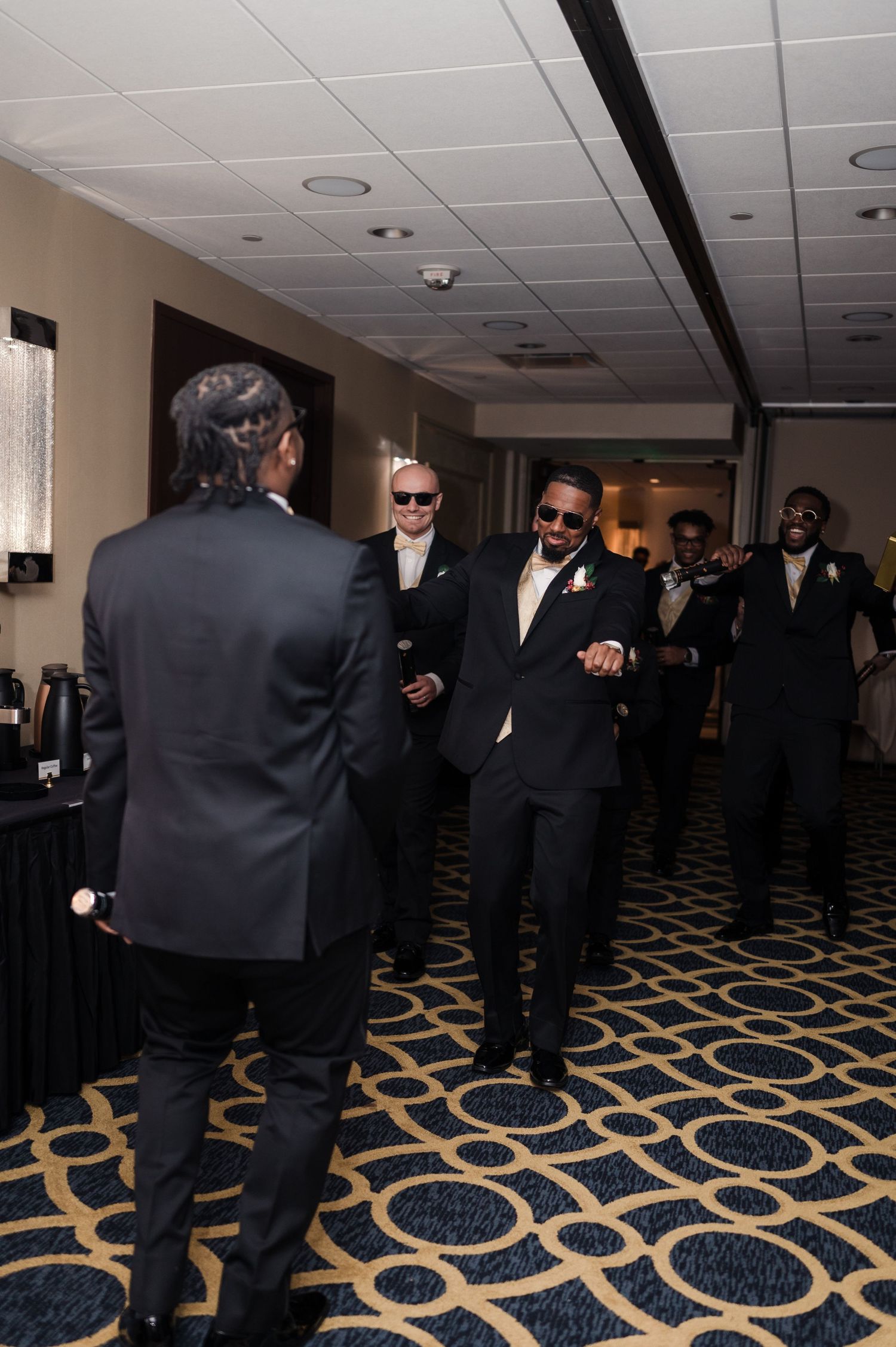Groomsmen in black suits and sunglasses pose together in a hallway with geometric patterned carpet.