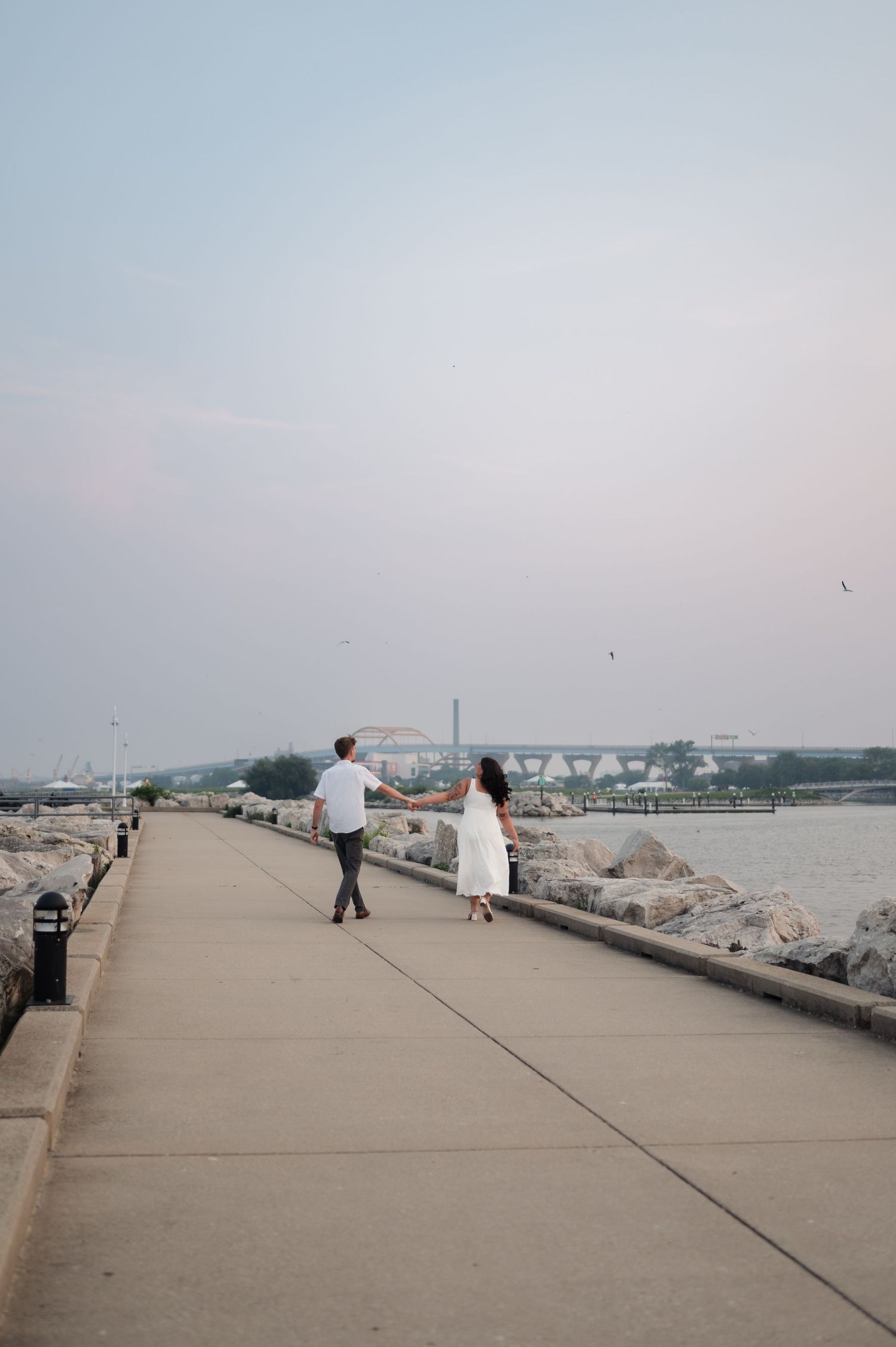 Two people walking along a concrete breakwater path by the water on a cloudy day with rocks lining the shoreline.