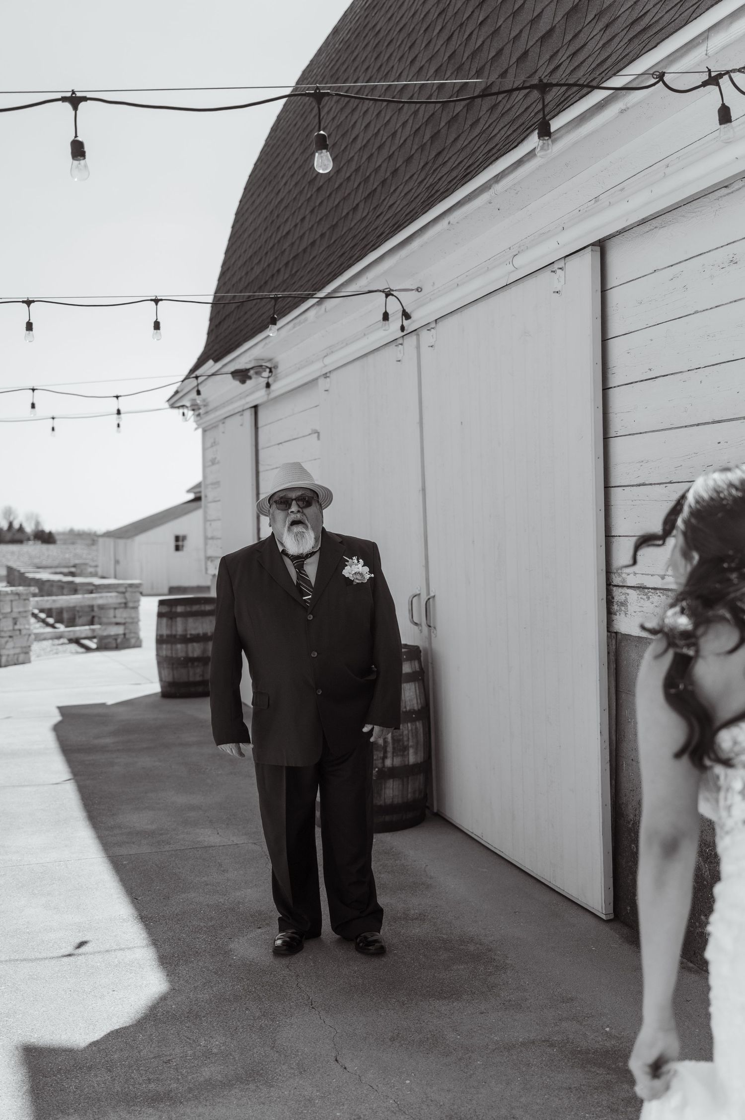 A black and white photo showing someone in a formal black suit standing next to a white barn with string lights above.