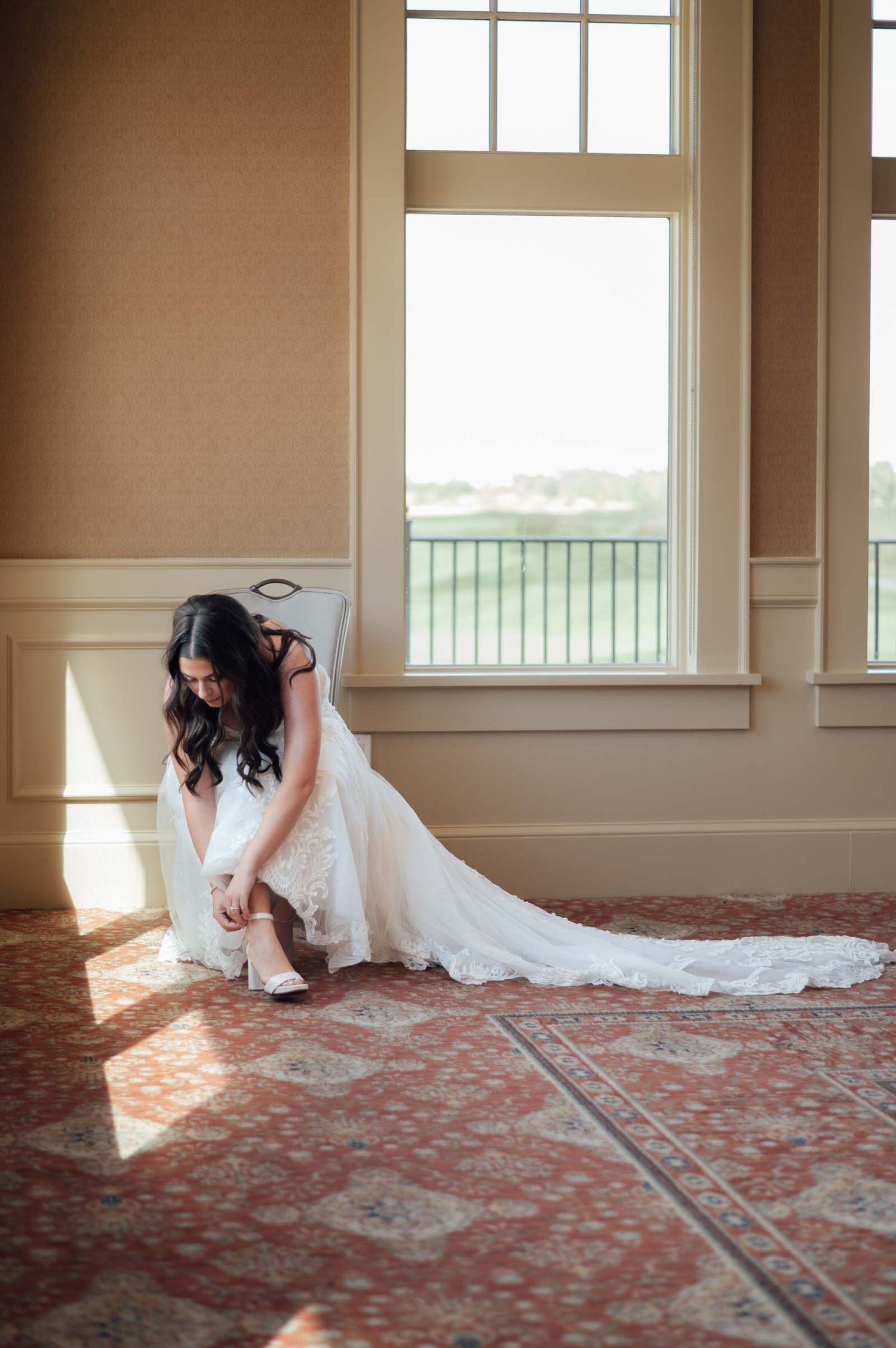 Bride in a white lace gown adjusting her shoe by a sunlit window on patterned carpet.