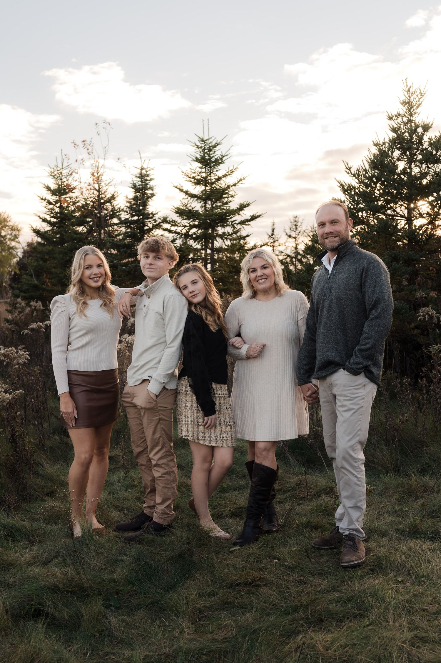 Family of five posing outdoors at sunset near evergreen trees in coordinated neutral fall outfits.