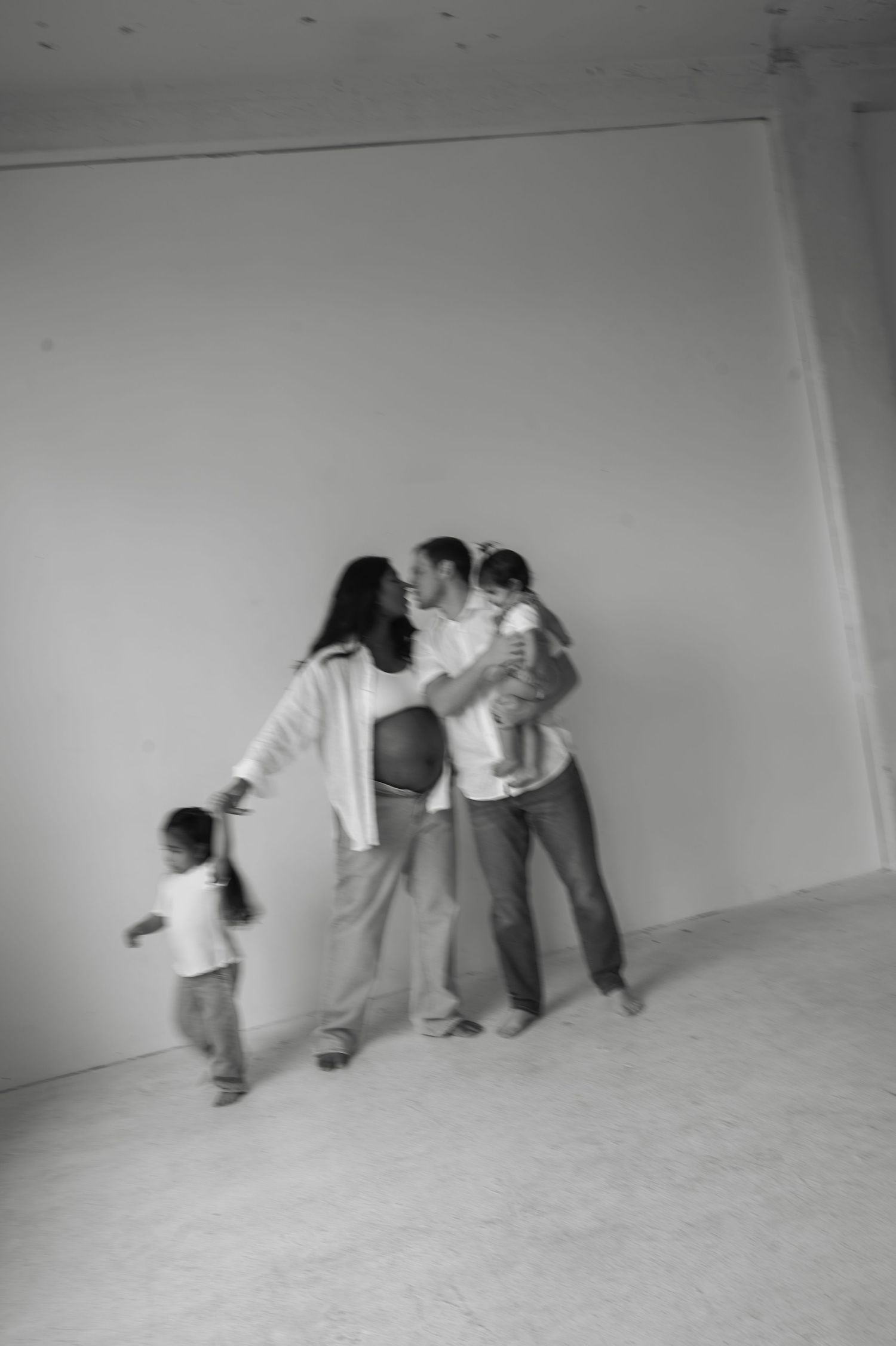 Black and white photo of family with pregnant mother, father, and two children against white wall.