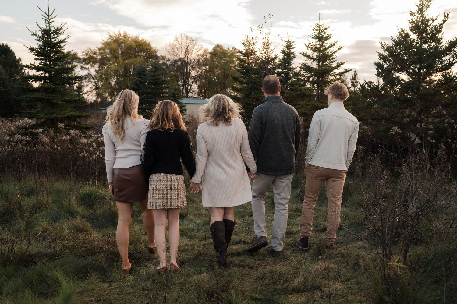 Five family members walk hand-in-hand through a pine tree farm at golden hour.