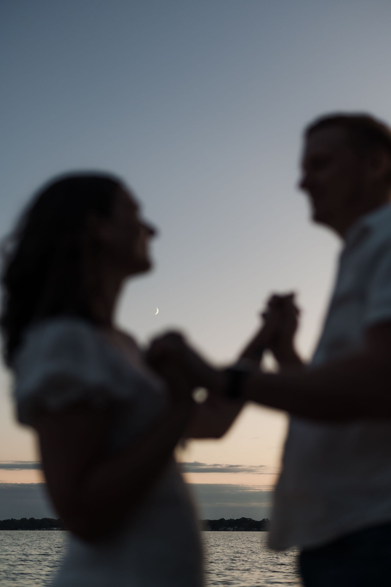 Silhouetted couple dances together at twilight by the water with a crescent moon visible in sky.