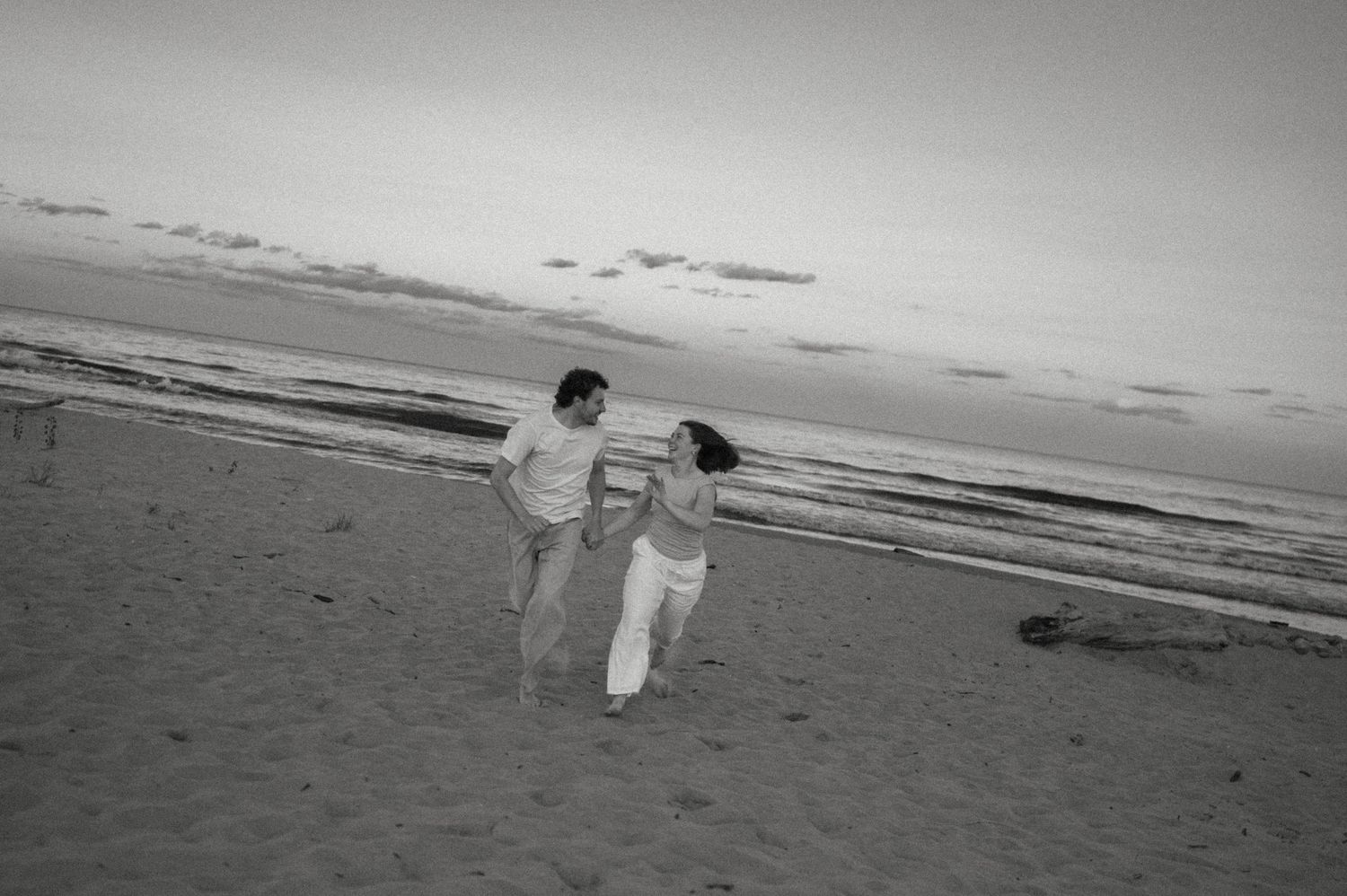 Black and white photo of a couple holding hands and walking on a sandy beach at sunset.