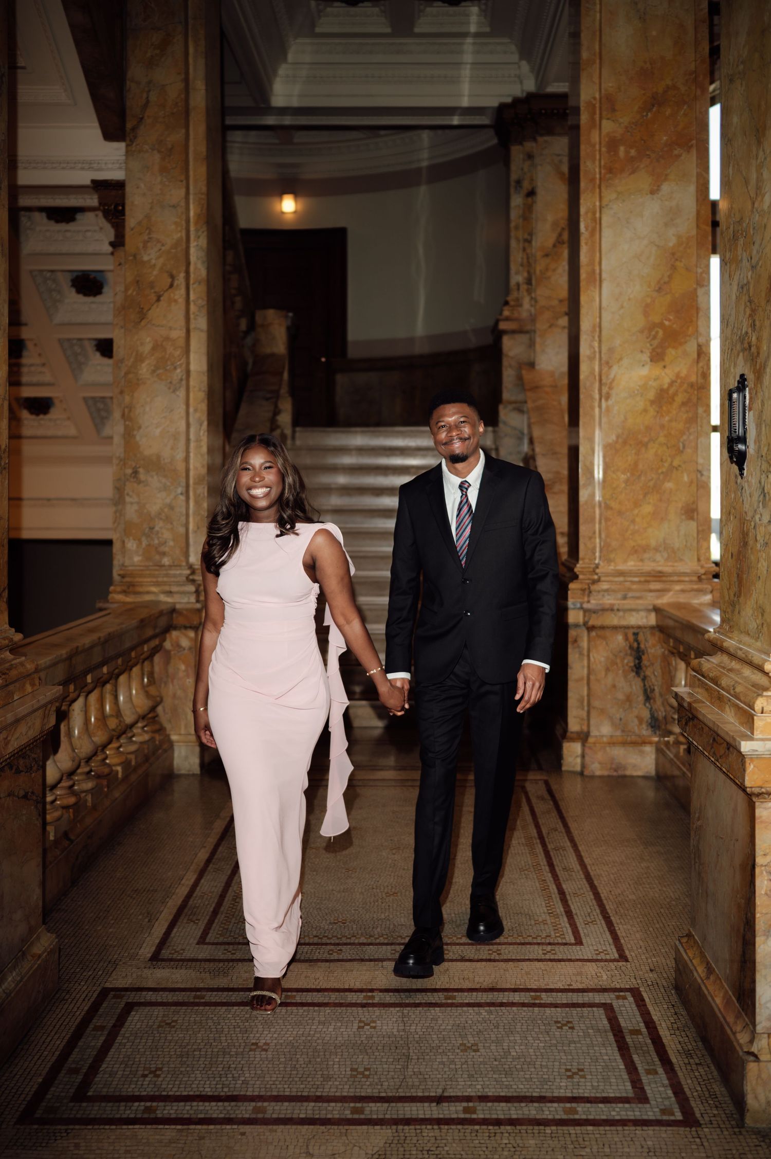 Elegant couple holding hands walking through a grand marble hallway, woman in white gown and man in black suit.