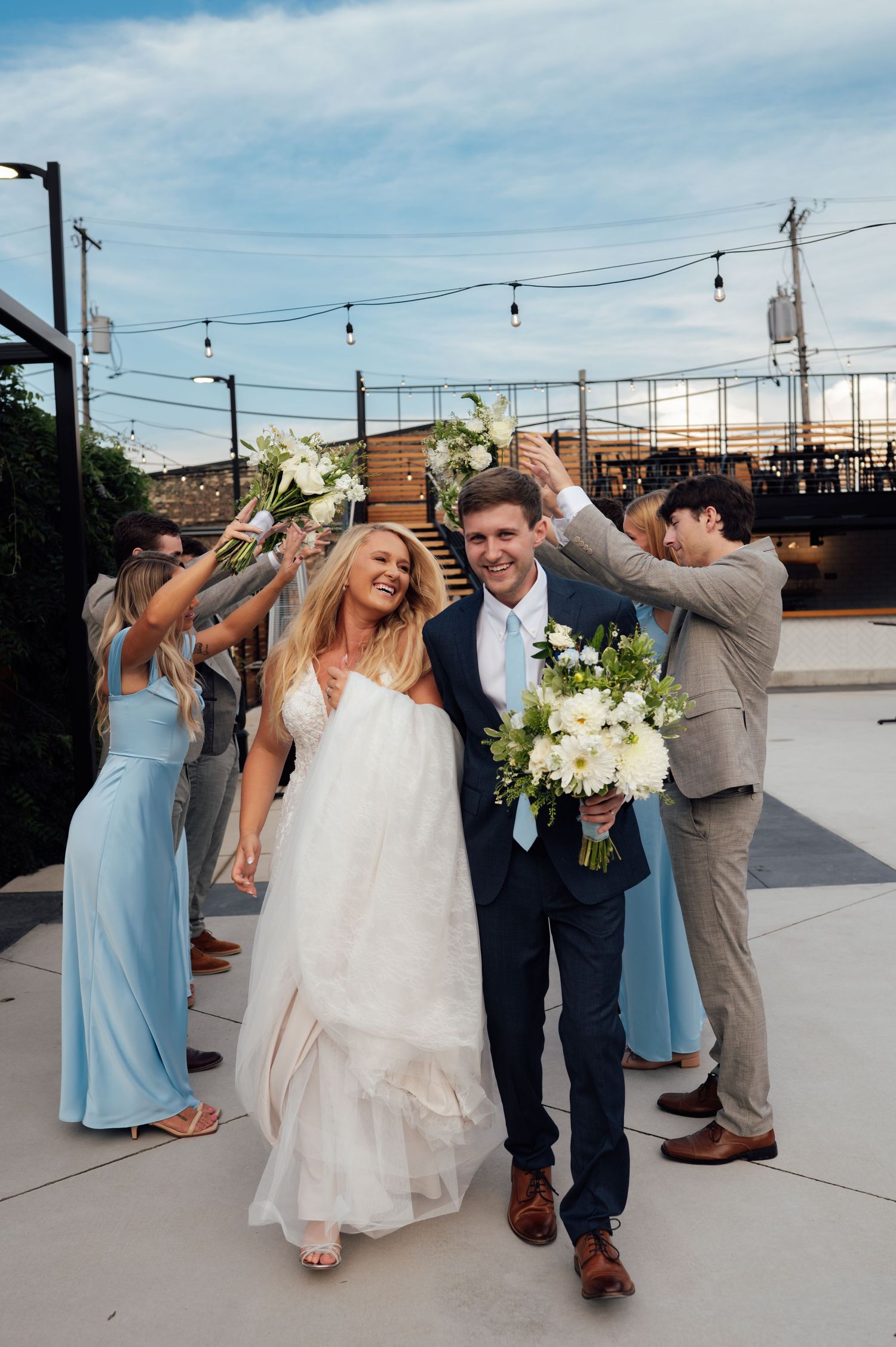Bride and groom exit wedding ceremony with bridal party in blue dresses and tan suits, holding white bouquets outdoors.