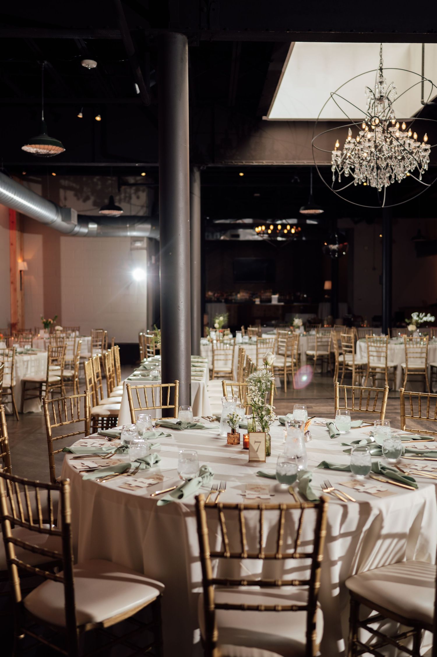 Elegant wedding reception venue with gold chiavari chairs, white round tables, crystal chandeliers, and soft floral centerpieces.