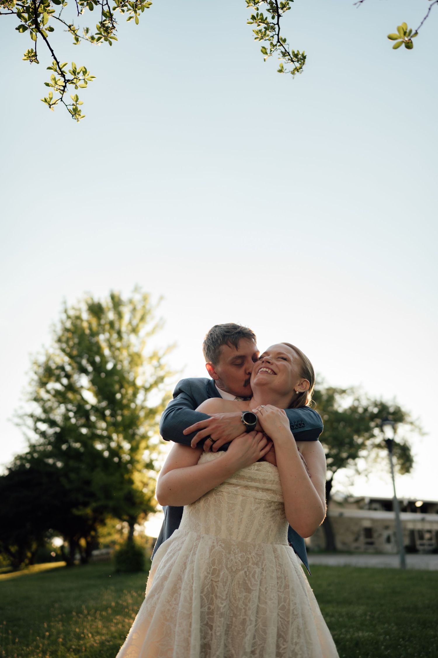 Groom embracing bride from behind in lace wedding dress outdoors at golden hour, both smiling joyfully under blooming tree branches.