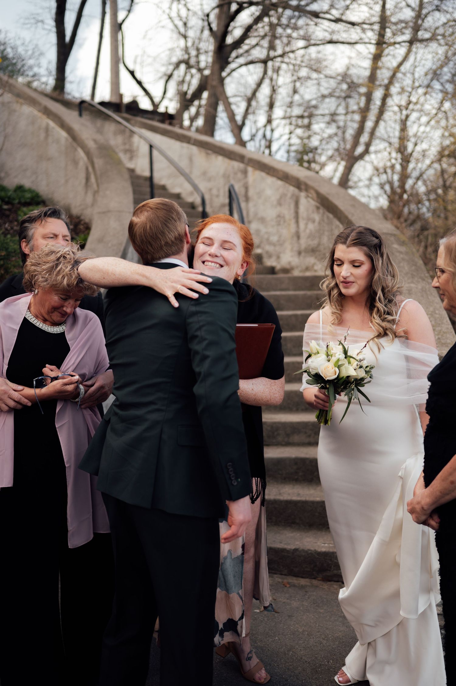 Bride in white gown holds bouquet as groom hugs redhead guest at outdoor wedding ceremony near stone steps.