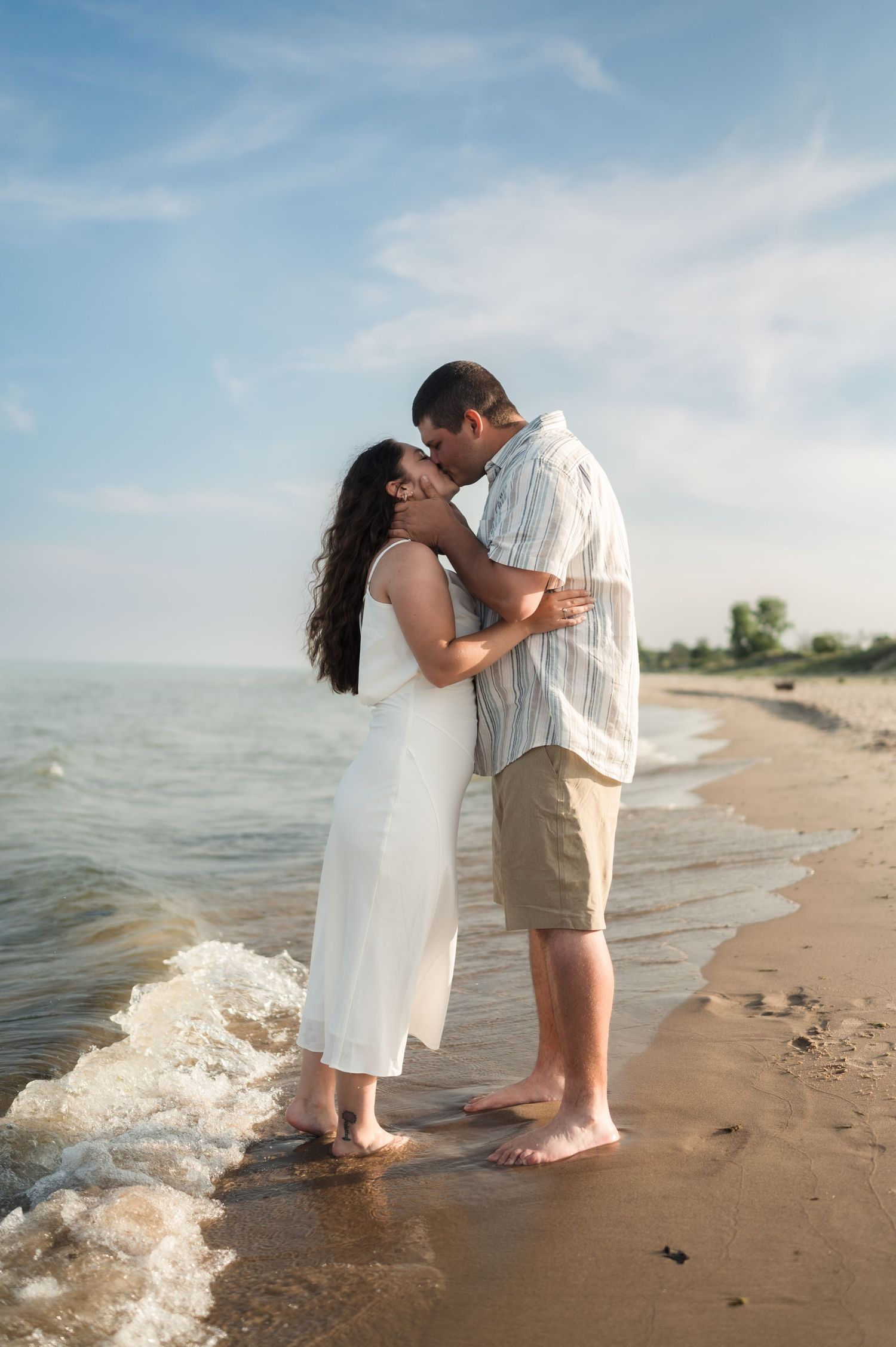A couple shares a romantic moment while standing barefoot on a sandy beach at the water's edge.