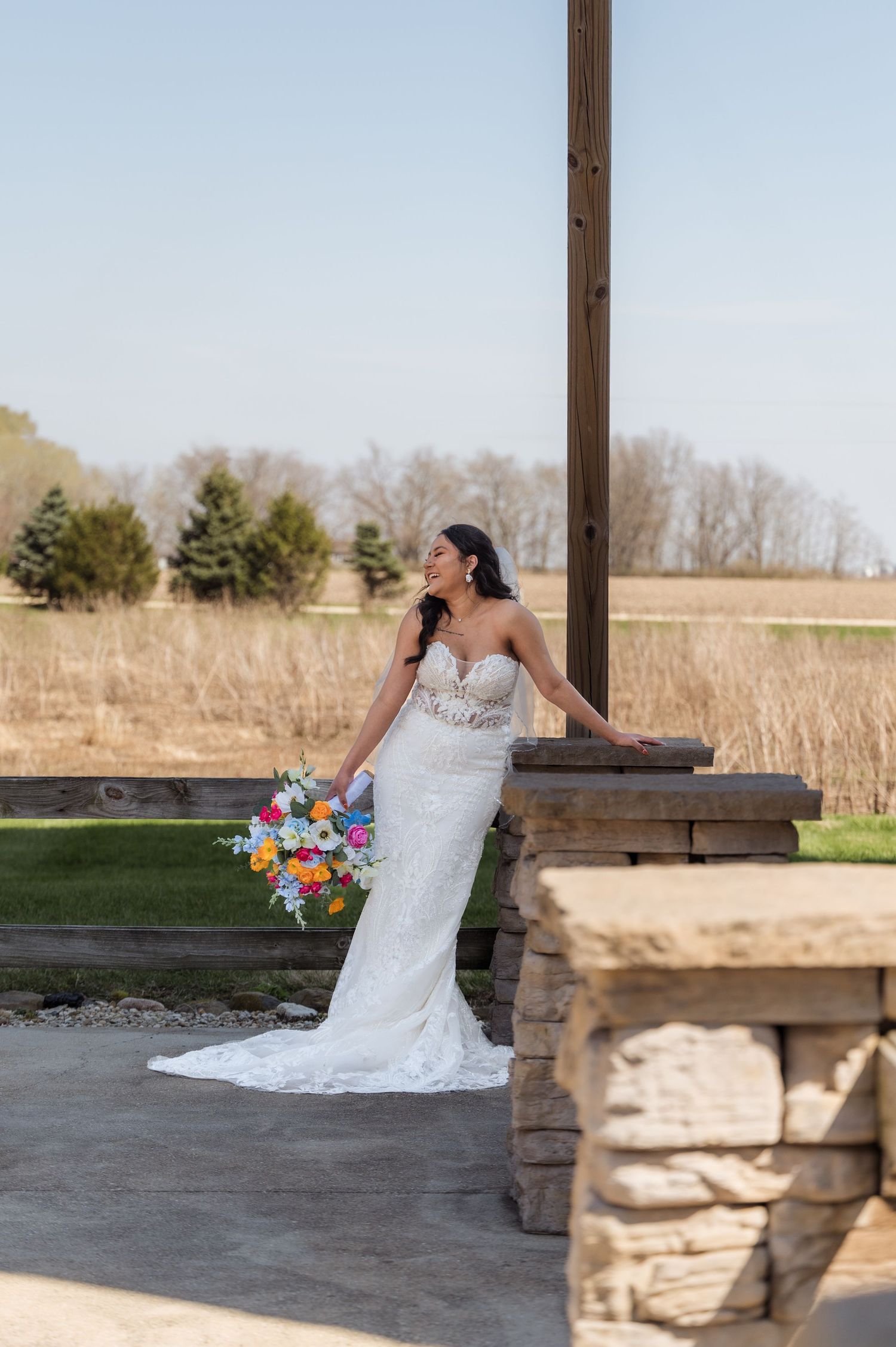 A bride in a fitted lace wedding dress poses with a colorful bouquet on a stone patio with rustic landscaping.