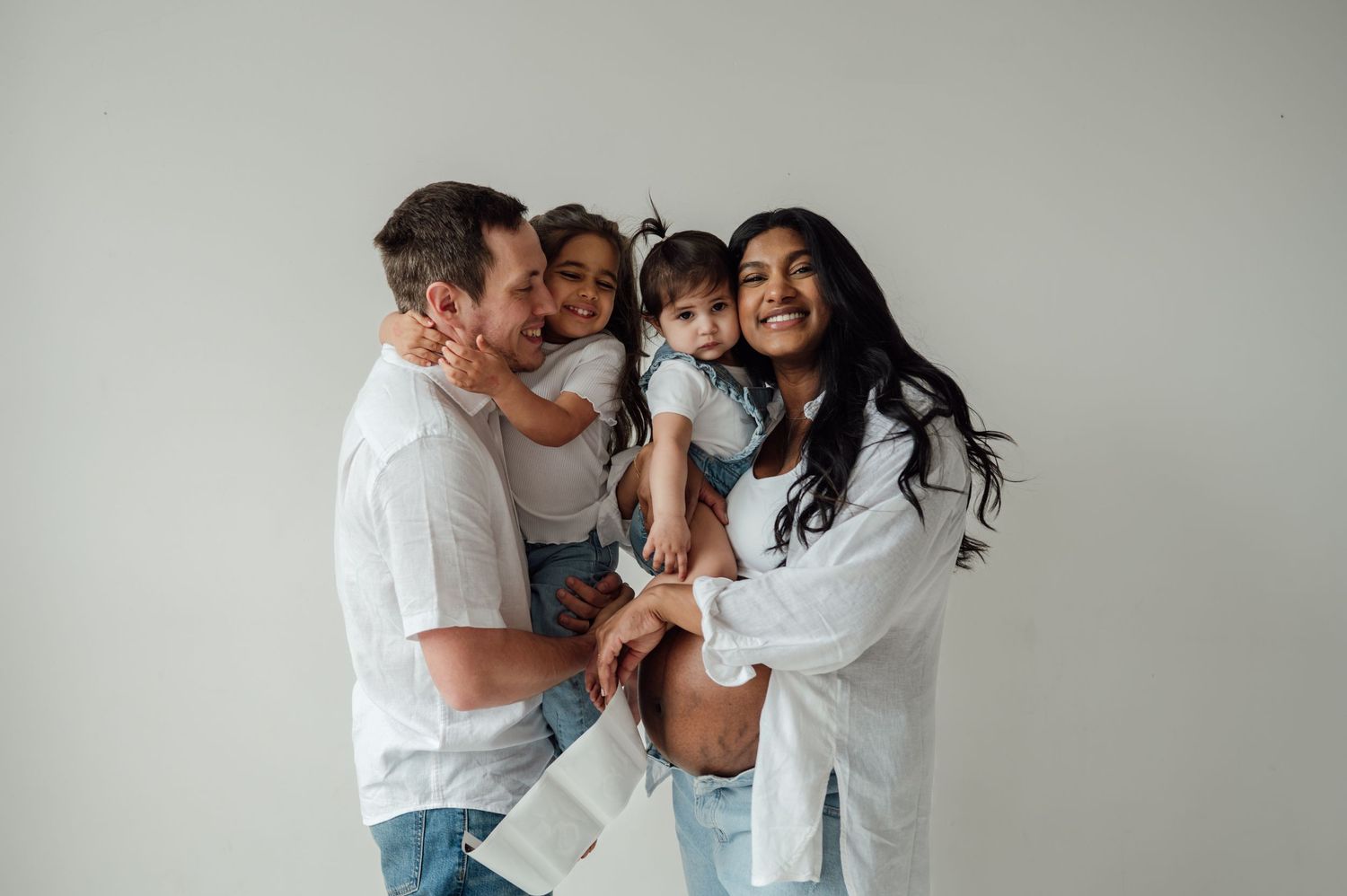 Happy family of four in white shirts and jeans laughing together against light gray background.
