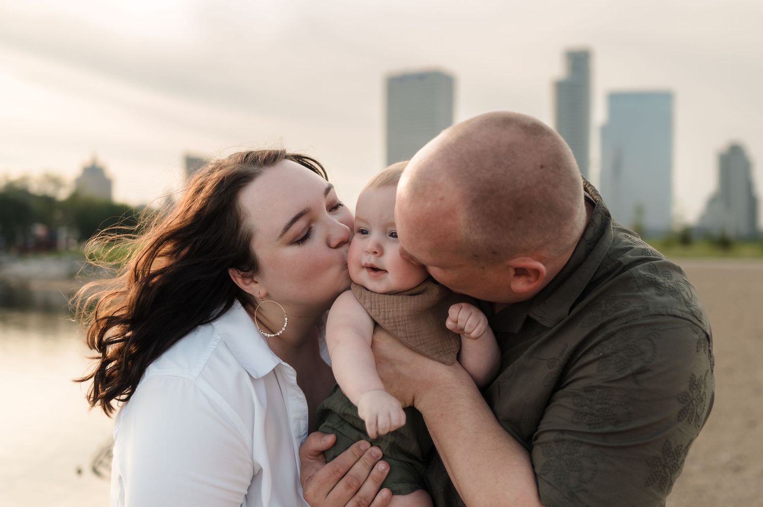Loving parents kissing their baby on the cheeks outdoors with a city skyline in the background.