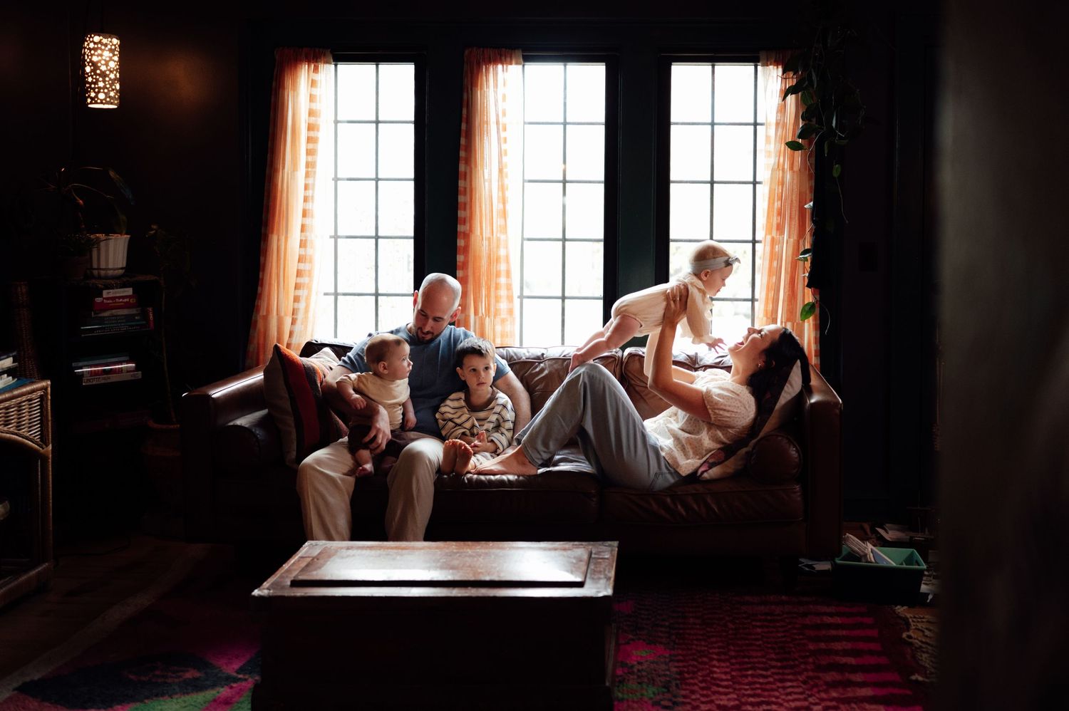 A family relaxes together on a leather couch in a warmly lit living room with large windows.