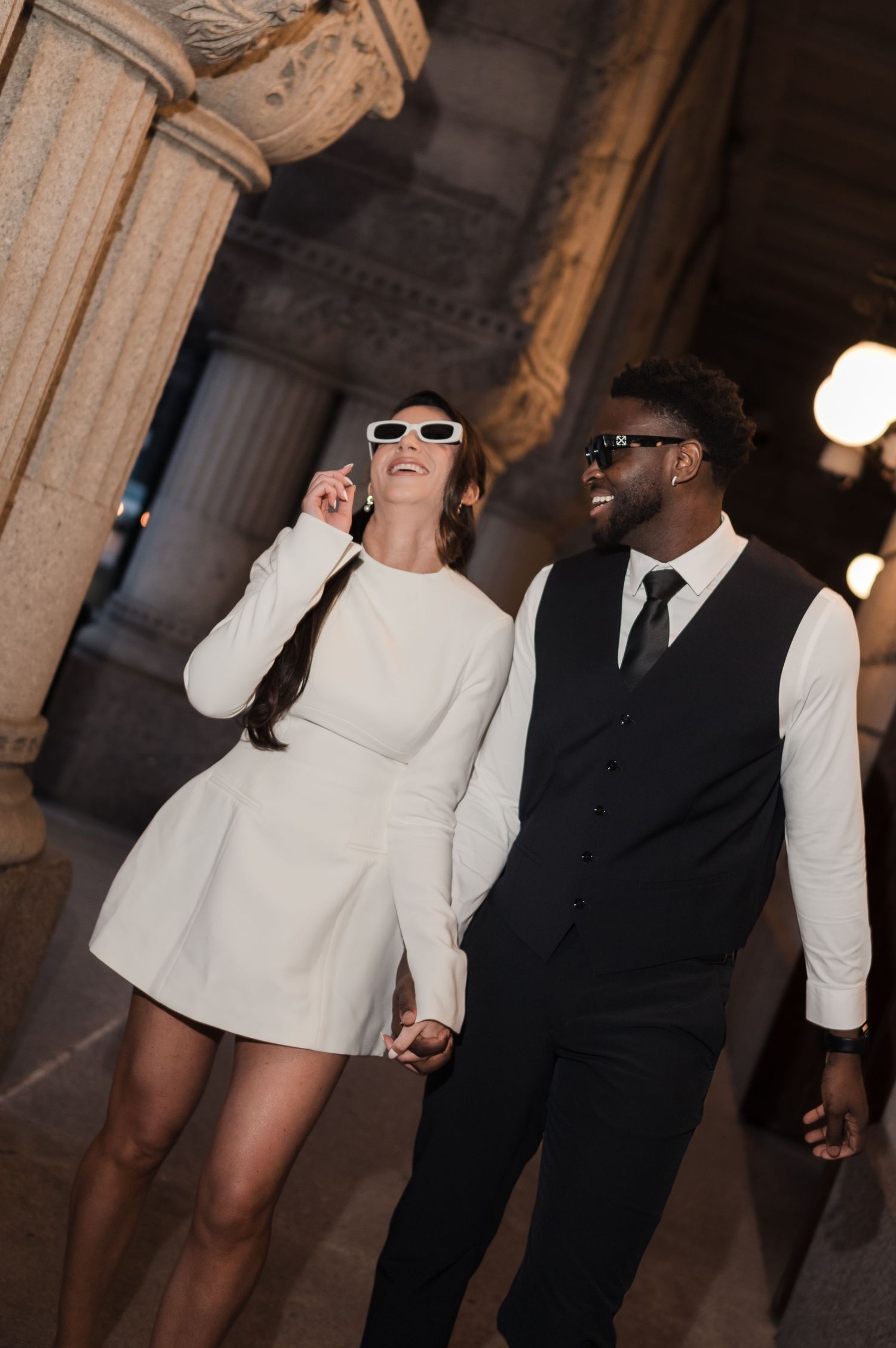 Stylish interracial couple holding hands and laughing while wearing sunglasses inside a grand architectural building.