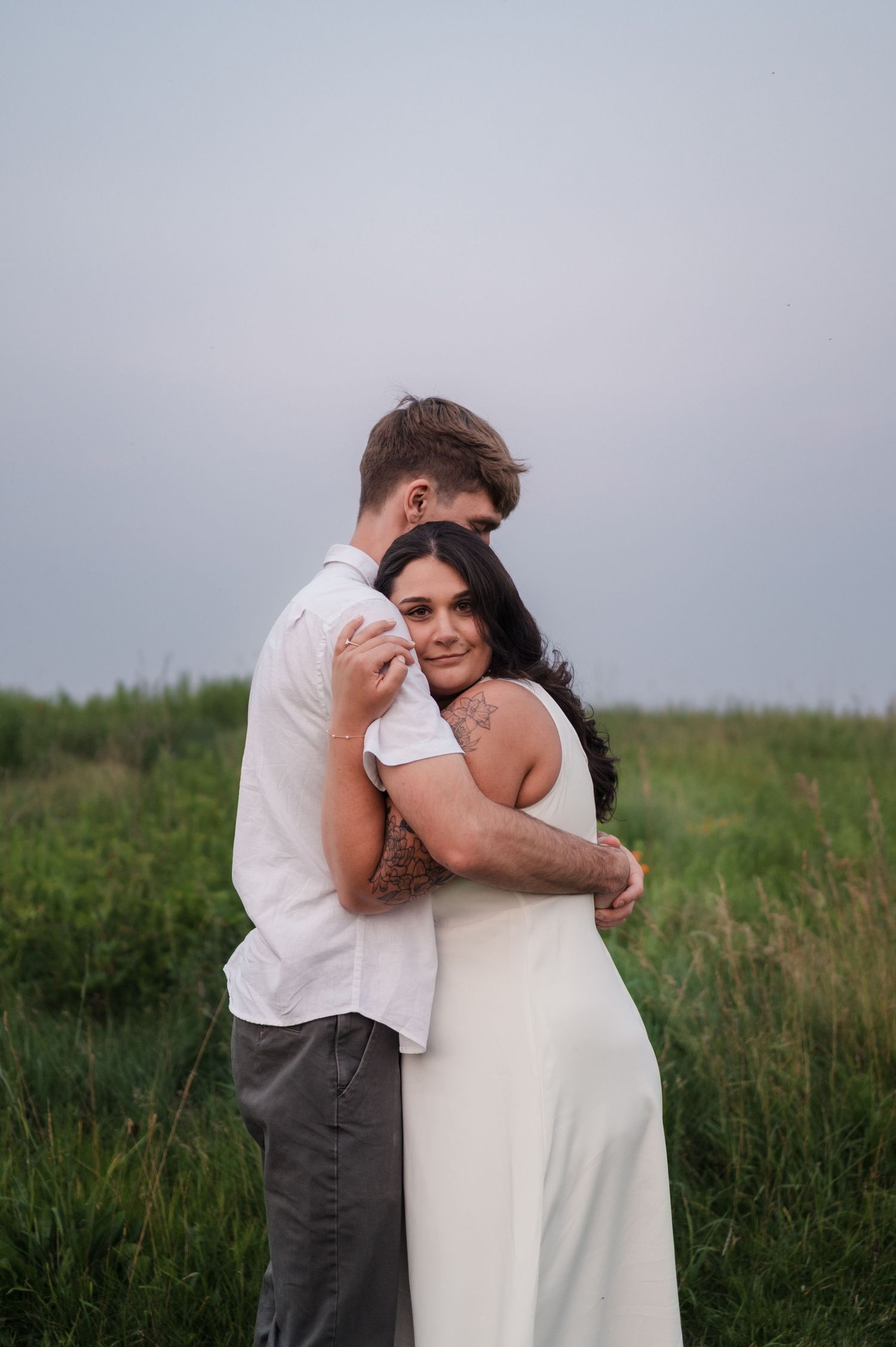 Woman with tattoos hugging her partner from behind in a green field during golden hour.