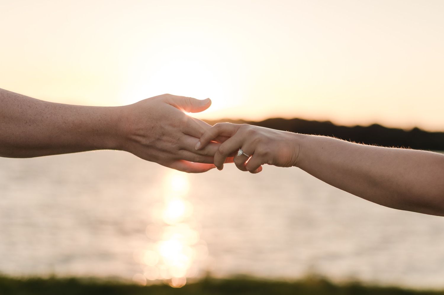Close-up of couple gently holding hands with sparkling sunset water reflecting in the background.