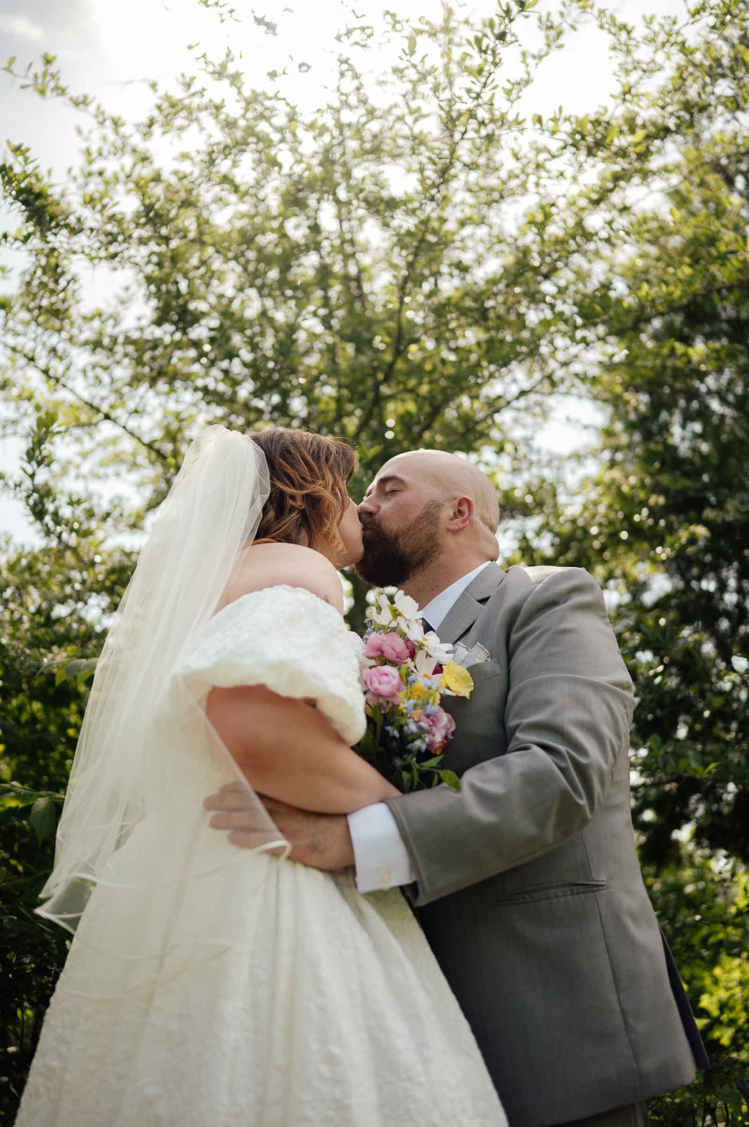 Bride and groom kissing under wedding veil surrounded by green trees outdoors.