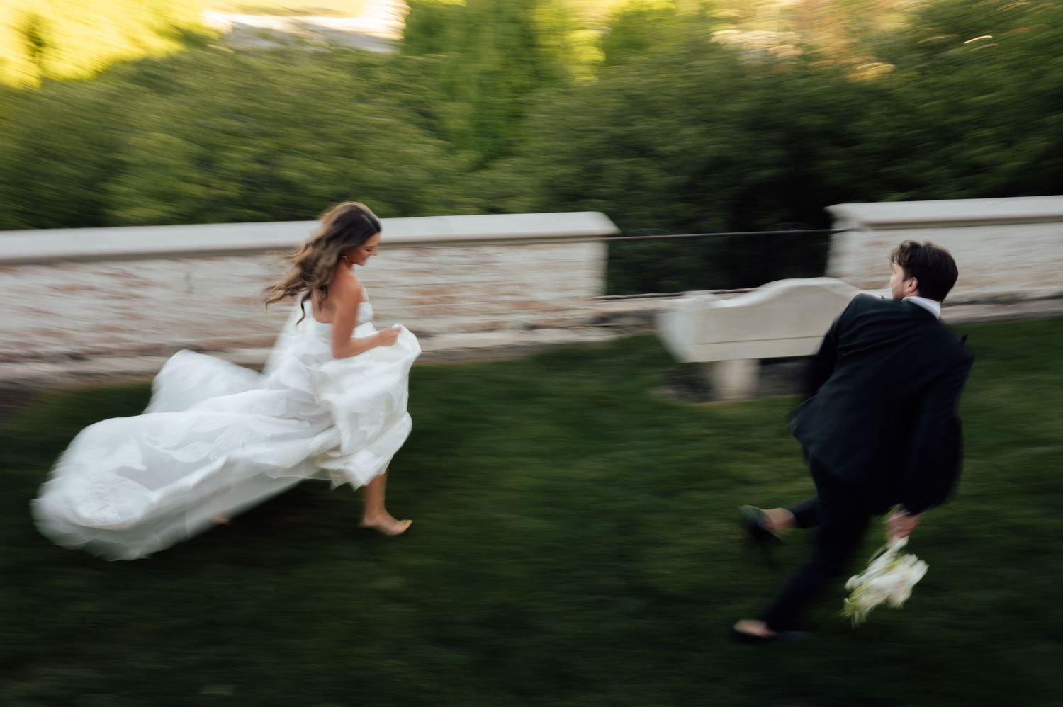 Bride and groom run playfully through lush green garden in a joyful motion-blurred wedding photo.