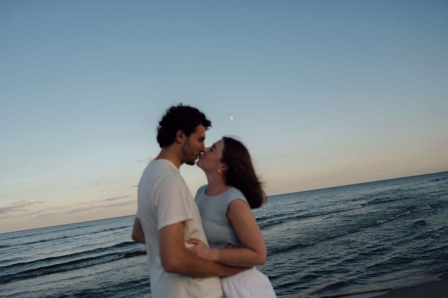 Romantic couple sharing a tender moment on a beach at dusk with ocean waves in the background.