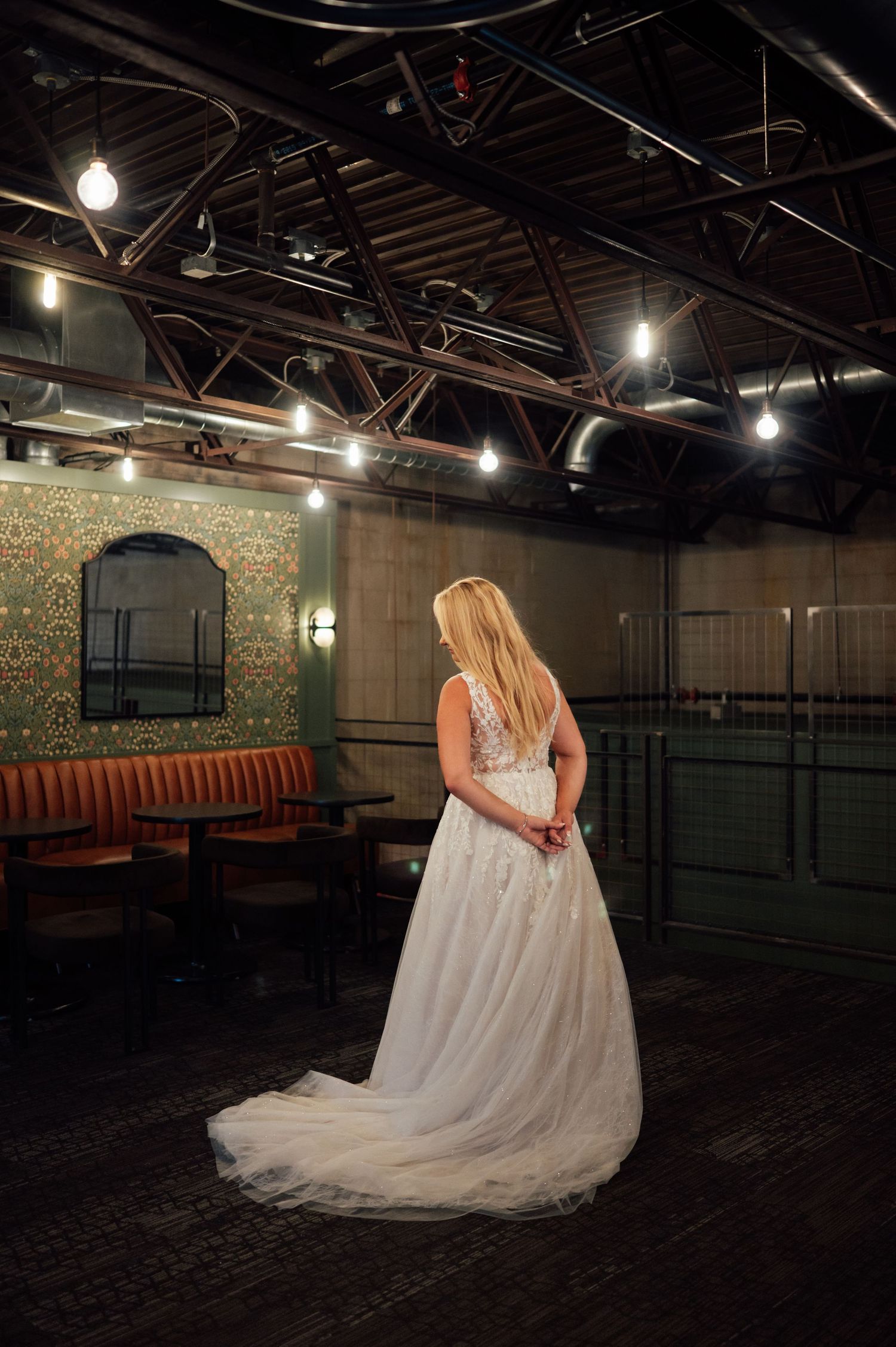Bride in elegant white tulle wedding gown with lace bodice stands in a vintage venue with warm Edison bulb lighting.