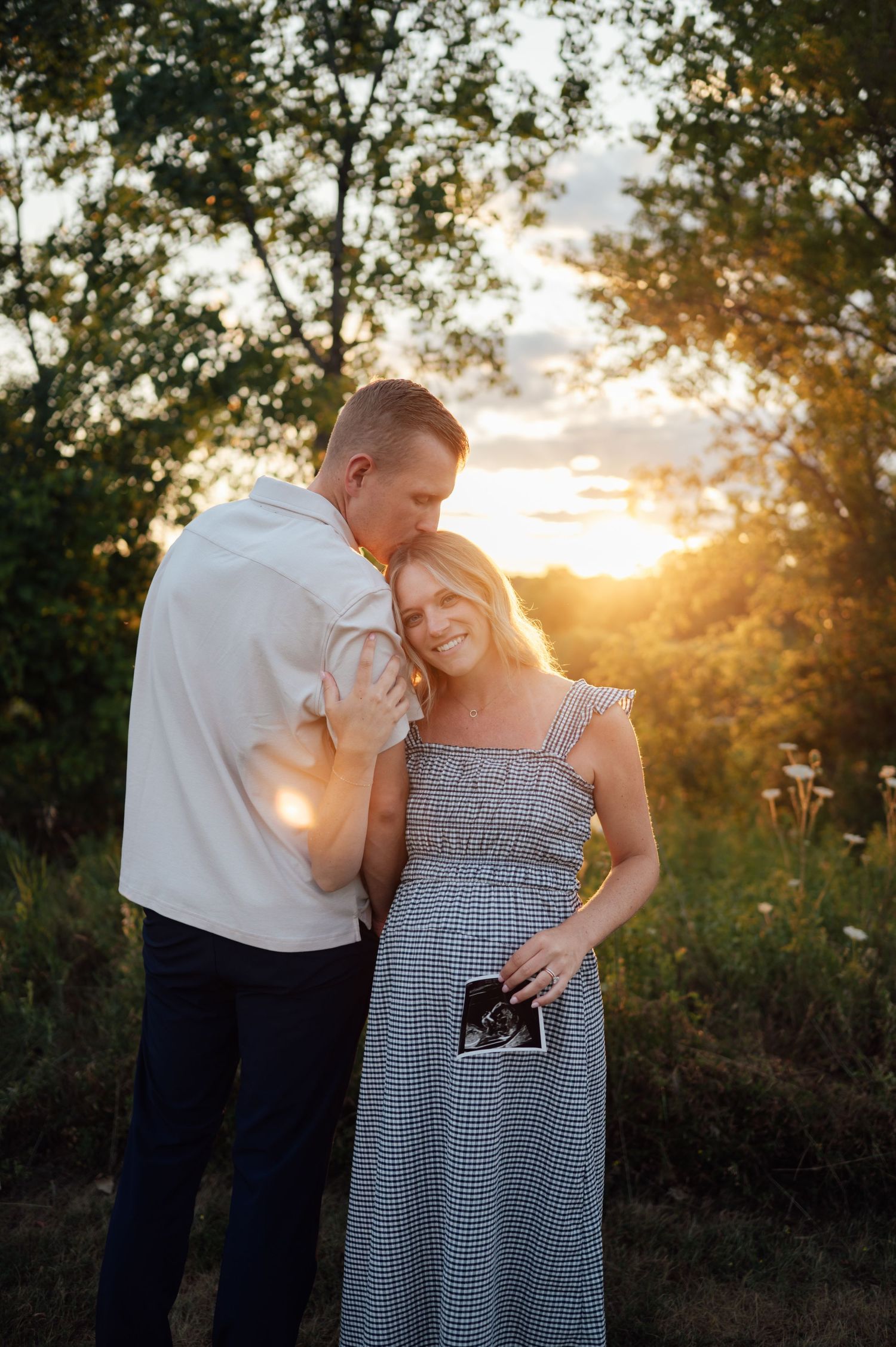 Couple sharing a tender moment at golden hour, woman holding ultrasound photo, pregnancy announcement outdoors.