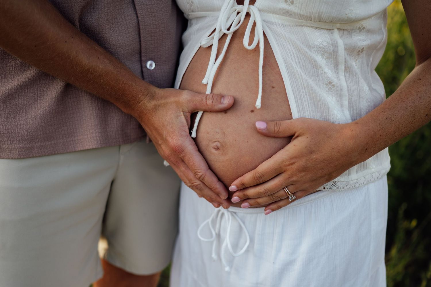 Pregnant woman in white dress with partner's hands forming heart shape on baby bump outdoors.