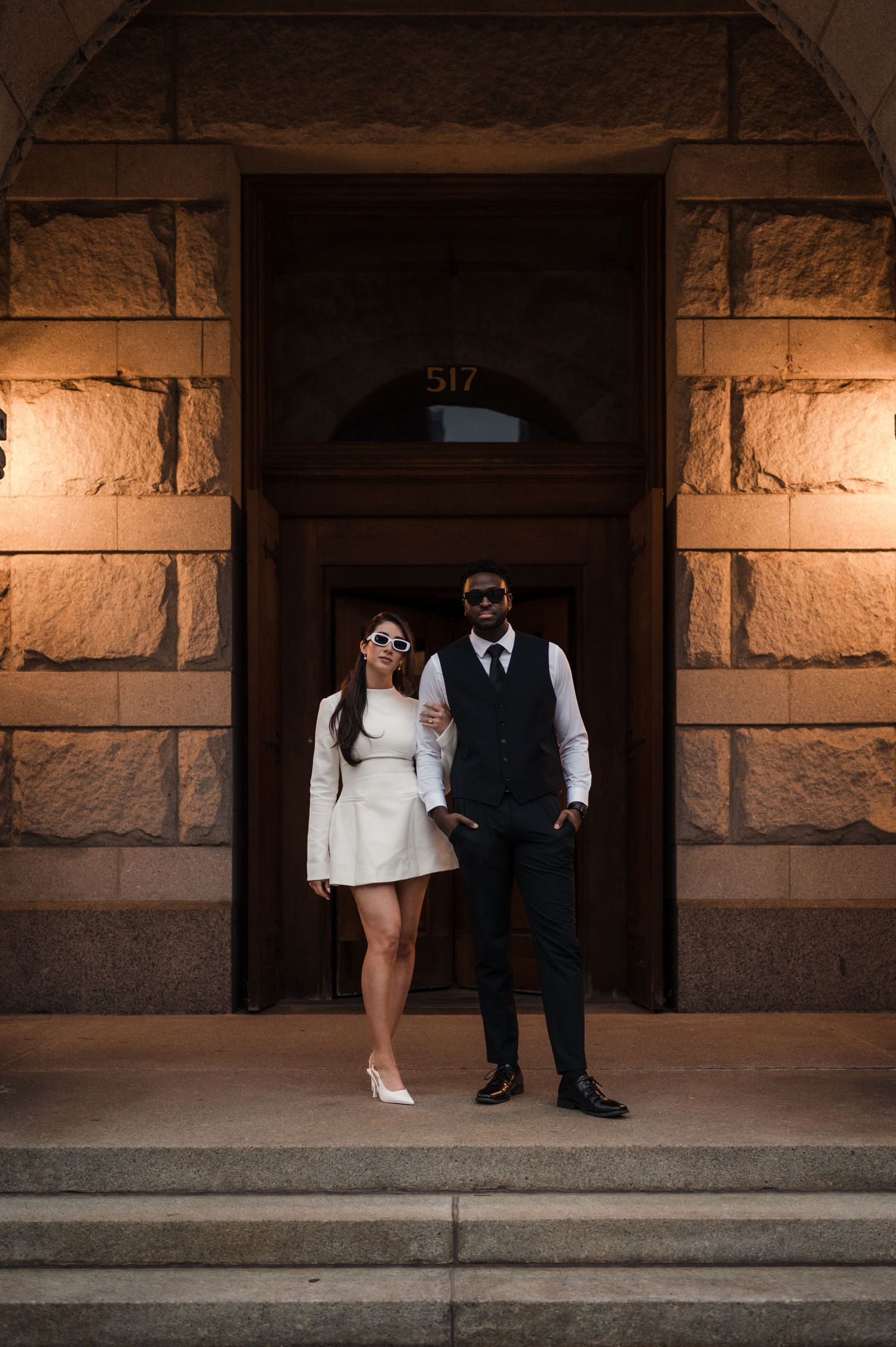 Stylish couple in white dress and black suit posing at stone building entrance numbered 517.