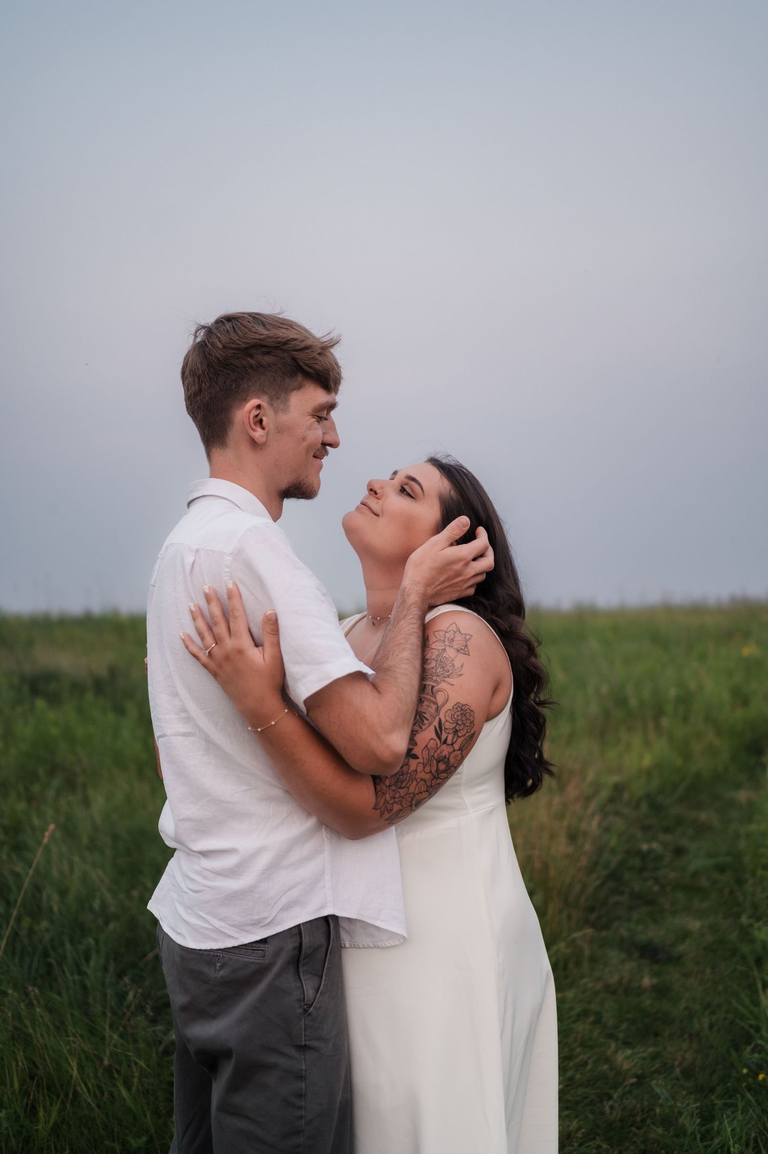 Couple embracing and gazing at each other in a lush green field under a soft sky.