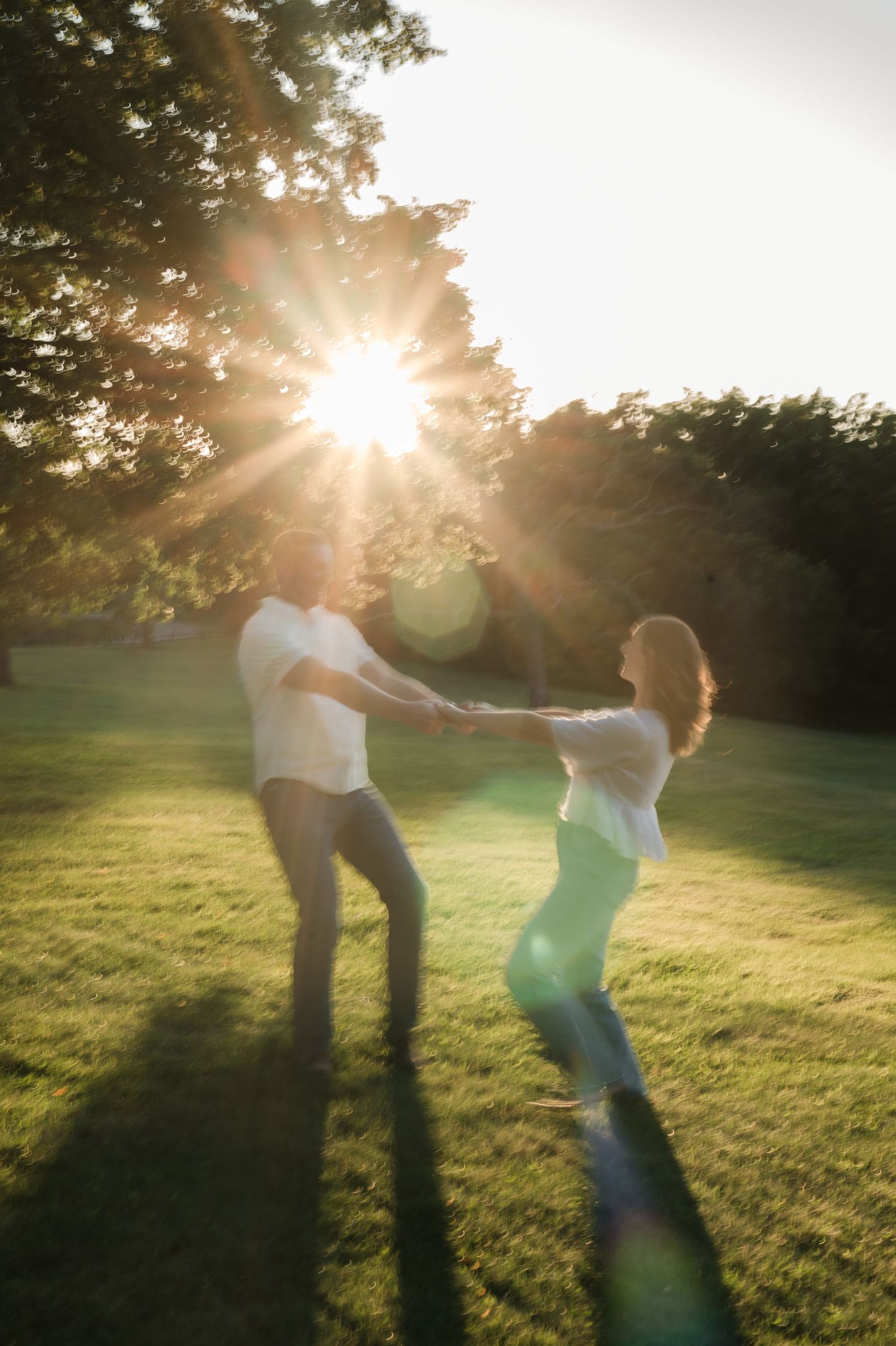 Couple spinning and dancing together in sunlit park with golden sun flare bursting through tall trees.