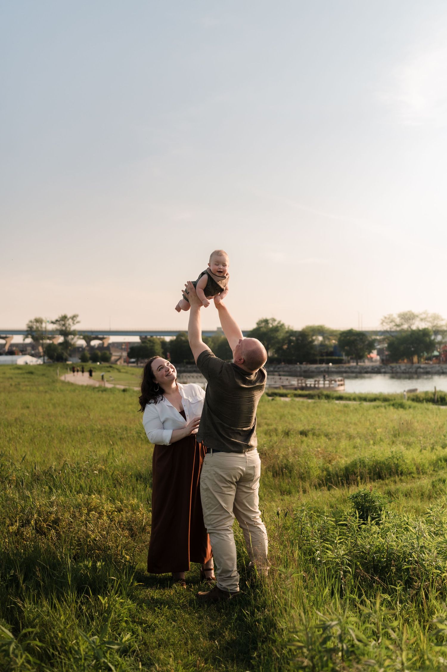 Joyful parents lifting their laughing baby in the air on a grassy field near a river at sunset.