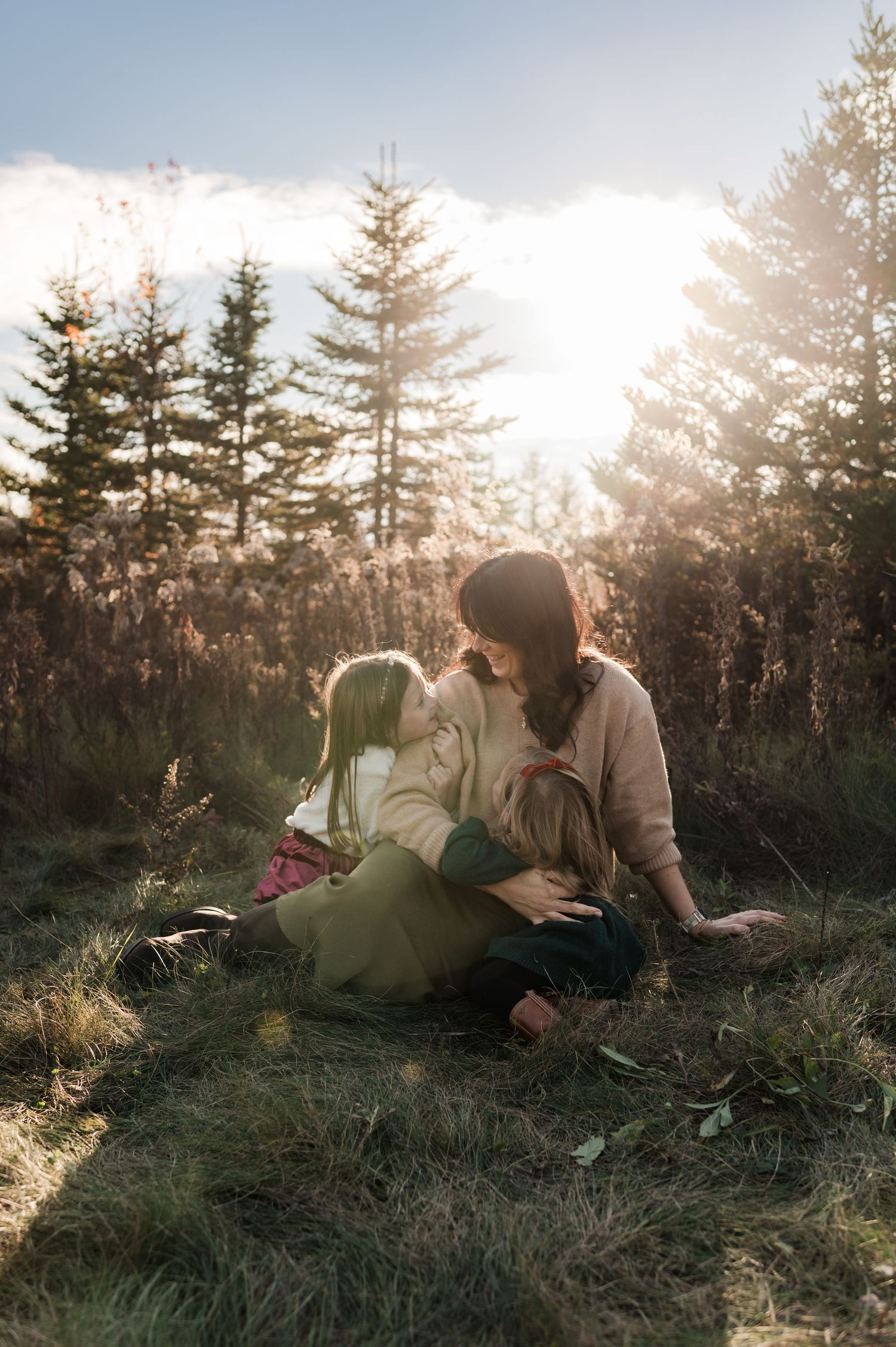 Mother sitting in golden sunlit grass embracing her two young daughters near pine trees.