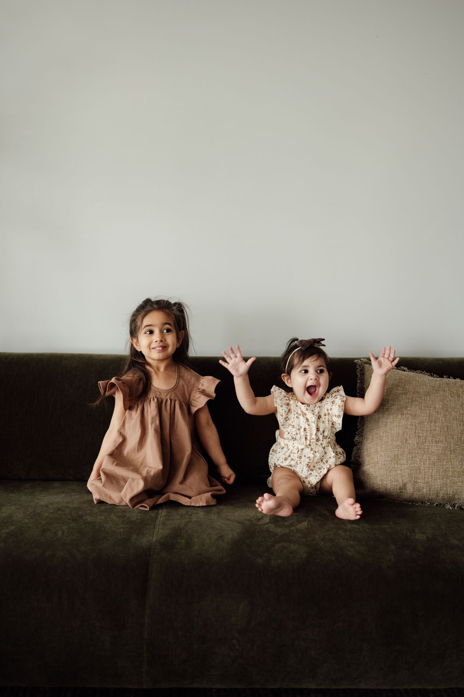 Two young sisters sitting on dark green sofa, older girl in brown dress, baby in floral romper.