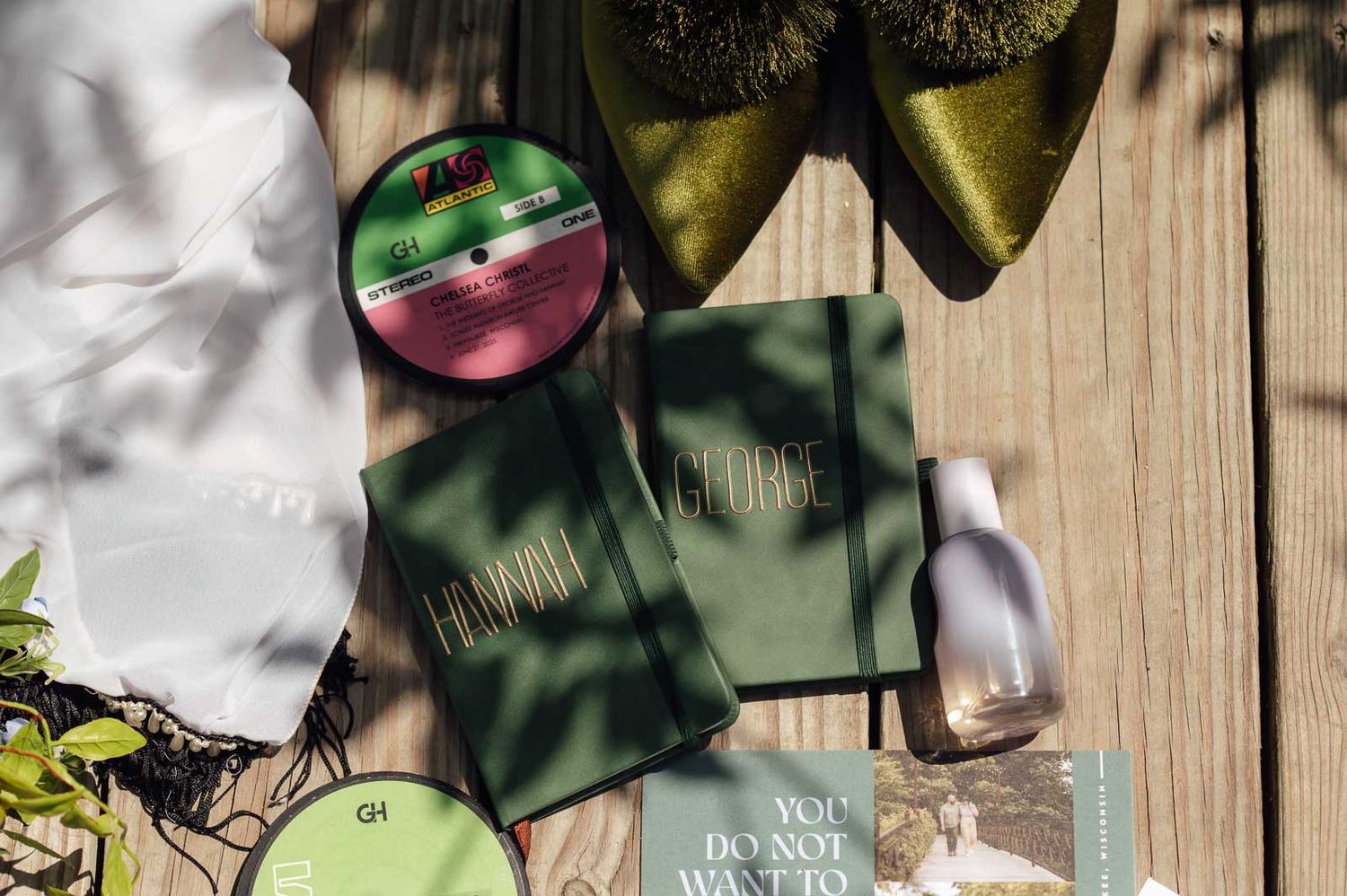 Flat lay of green wedding stationery, vinyl record, and beauty products on wooden surface.