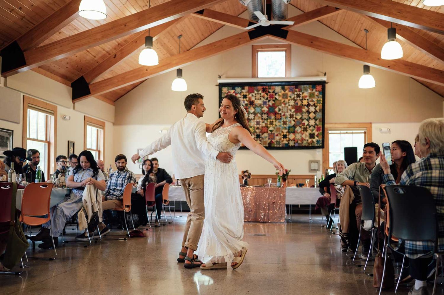 Bride and groom sharing first dance in rustic wood-beamed hall with guests applauding around them.