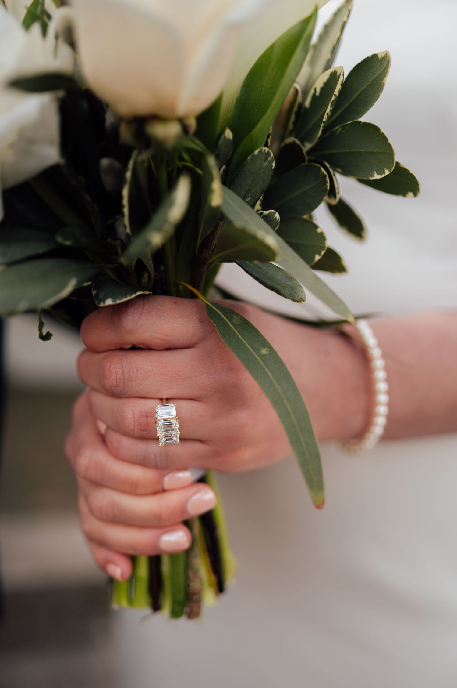 A close-up of a bride's hand wearing a diamond ring while holding a white rose and greenery bouquet.