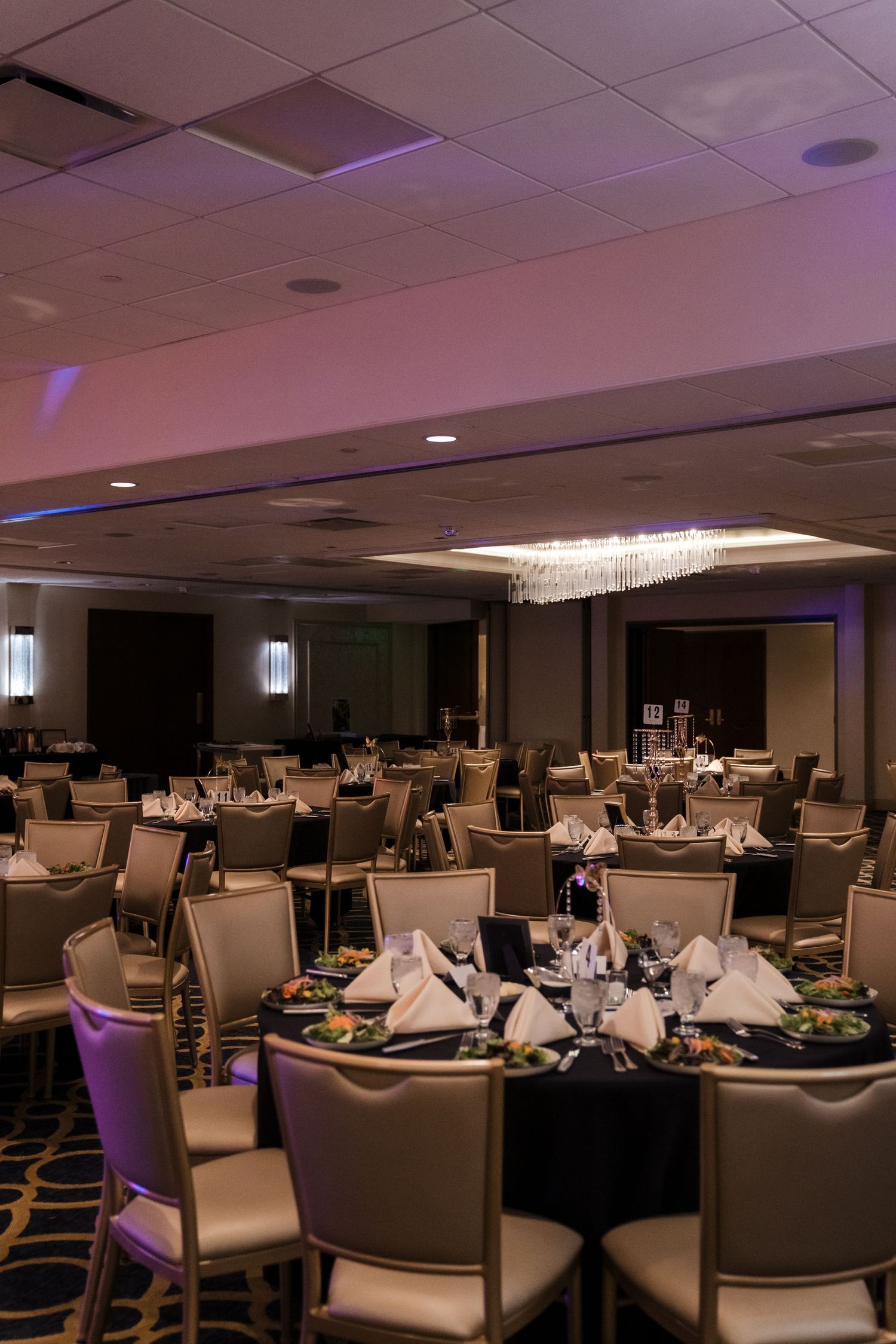 Elegant banquet hall setup with round tables and white chairs under crystal chandeliers for a formal event.