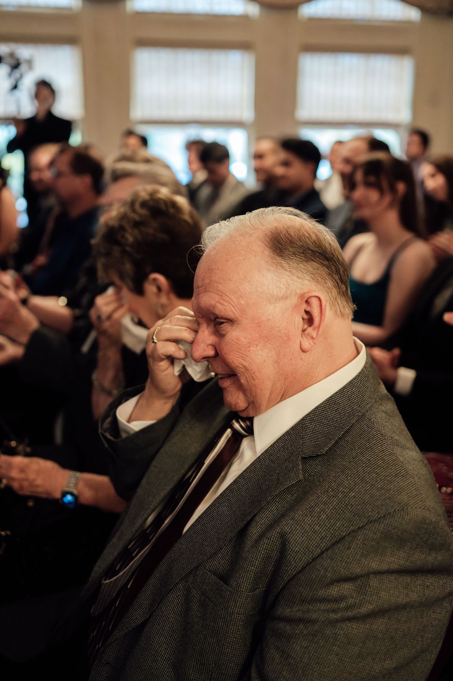Emotional older man in gray suit wiping tears at a crowded formal event or wedding ceremony.
