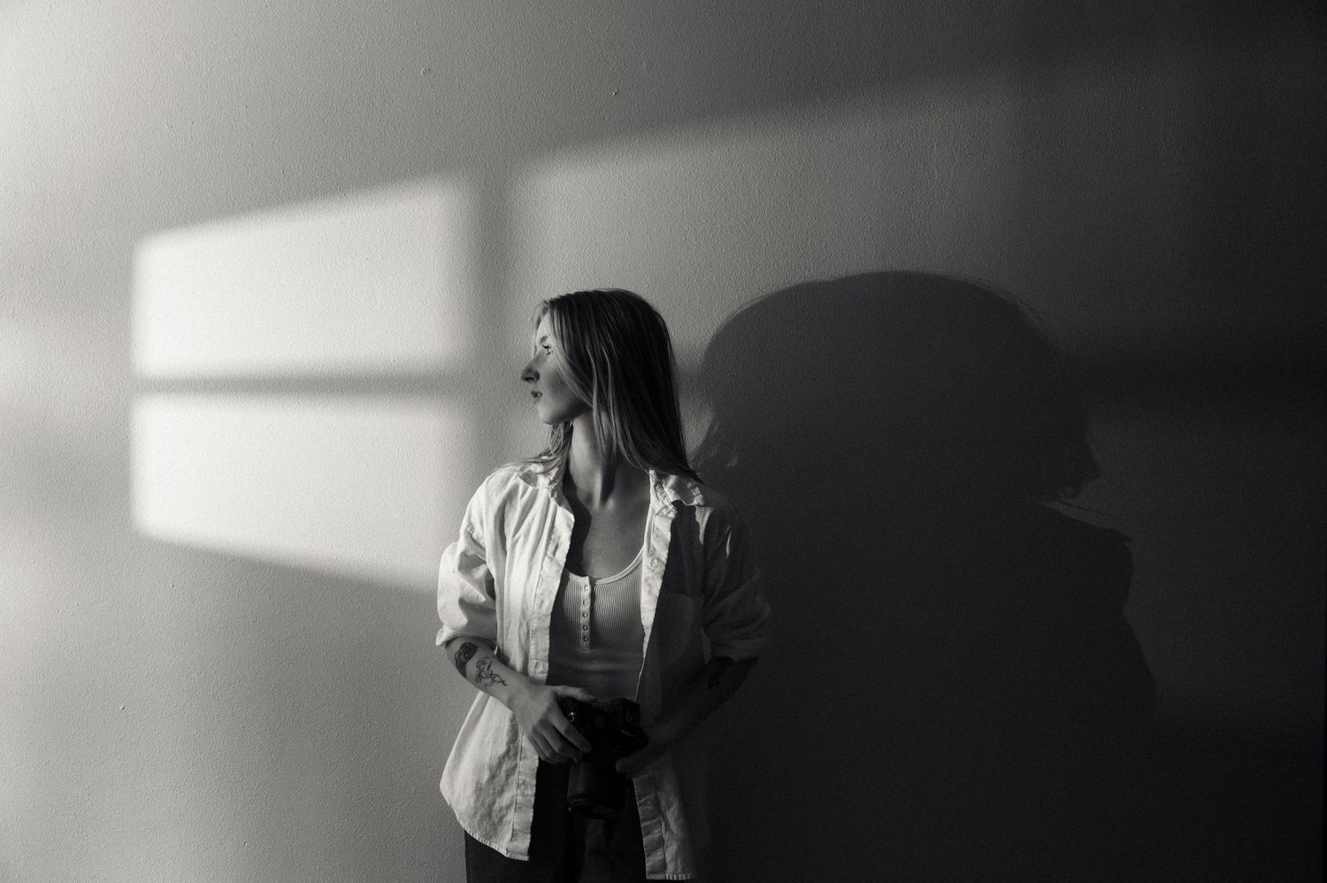 A woman in a white shirt stands against a wall with dramatic light and shadow in a black and white portrait.