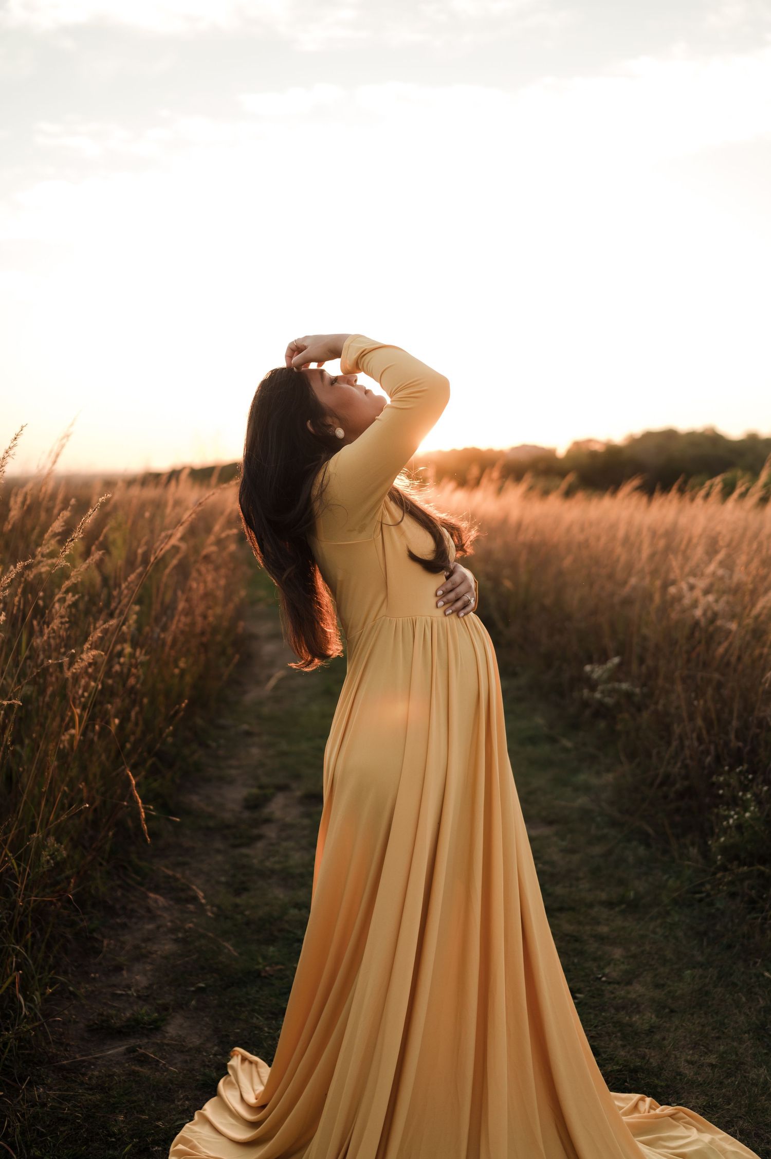Pregnant woman in flowing golden gown poses gracefully on a sunlit field path at sunset.